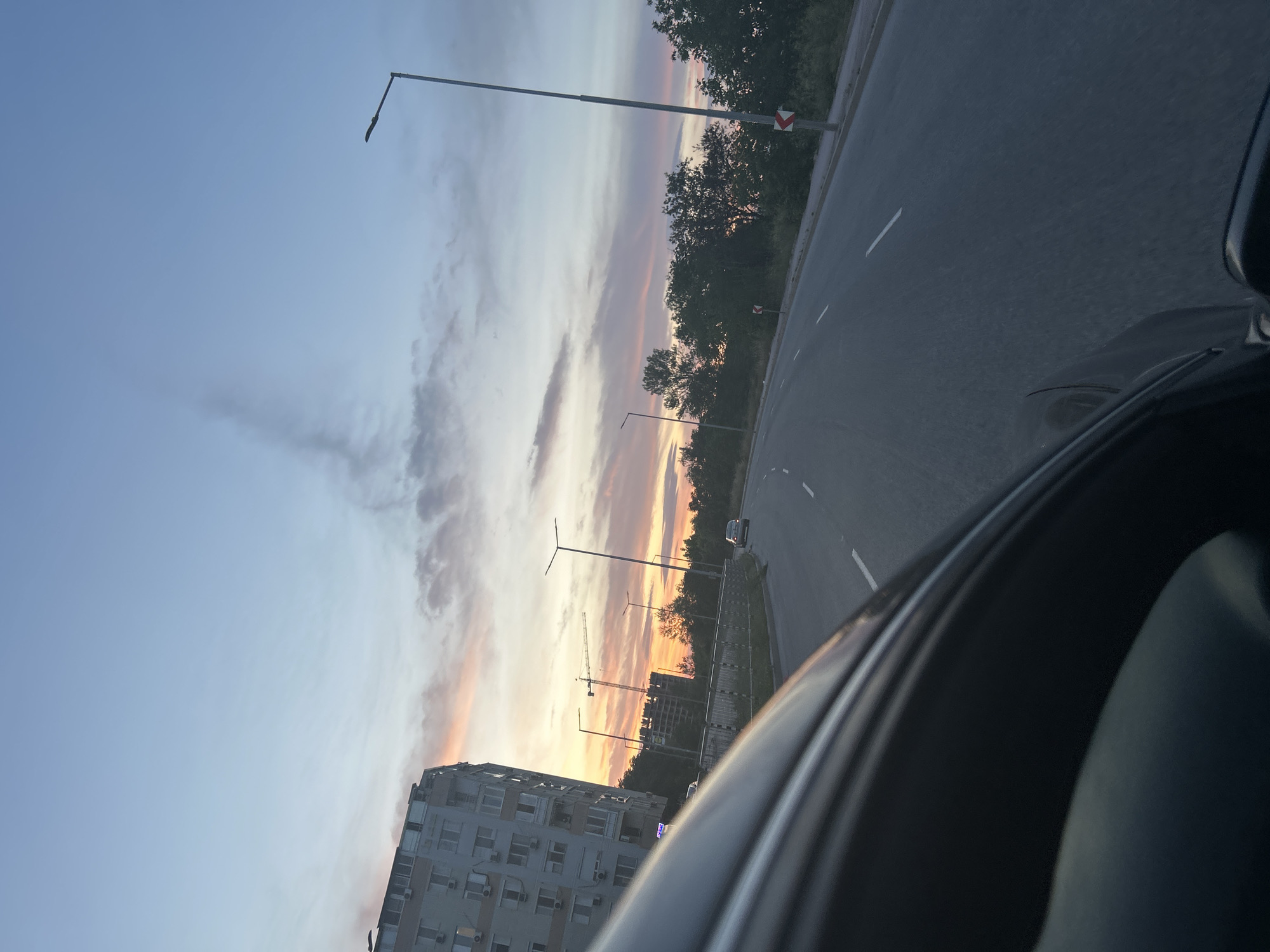 The image depicts a scene from Plovdiv, Bulgaria, at either sunrise or sunset. The perspective is from inside a moving vehicle on a multi-lane road. The road has white lane markings and a roadside area with trees and street lamps. In the distance, a building under construction with a crane is visible, along with other residential buildings and a clear, colorful sky.