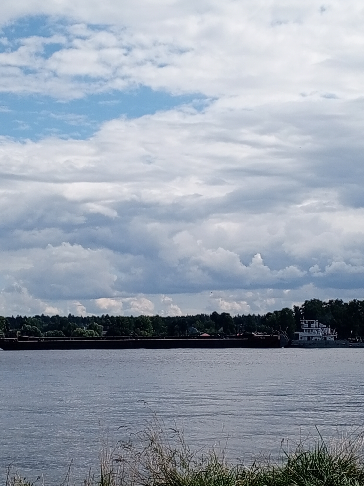 A long, dark barge is docked in the calm, blue waters of a river. The barge occupies the majority of the lower portion of the image. Behind the barge, a lush green forest lines the riverbank. A white building with multiple windows, possibly a boat house or a small structure, sits on the right side of the barge. The sky above is filled with a dramatic mix of white and gray clouds, with patches of blue sky peeking through. The foreground shows the tips of green grass swaying gently. The image appears to be taken from the shore, looking out over the river. The scene is set in Novoaleksandrovo, Russia, on a cloudy day.