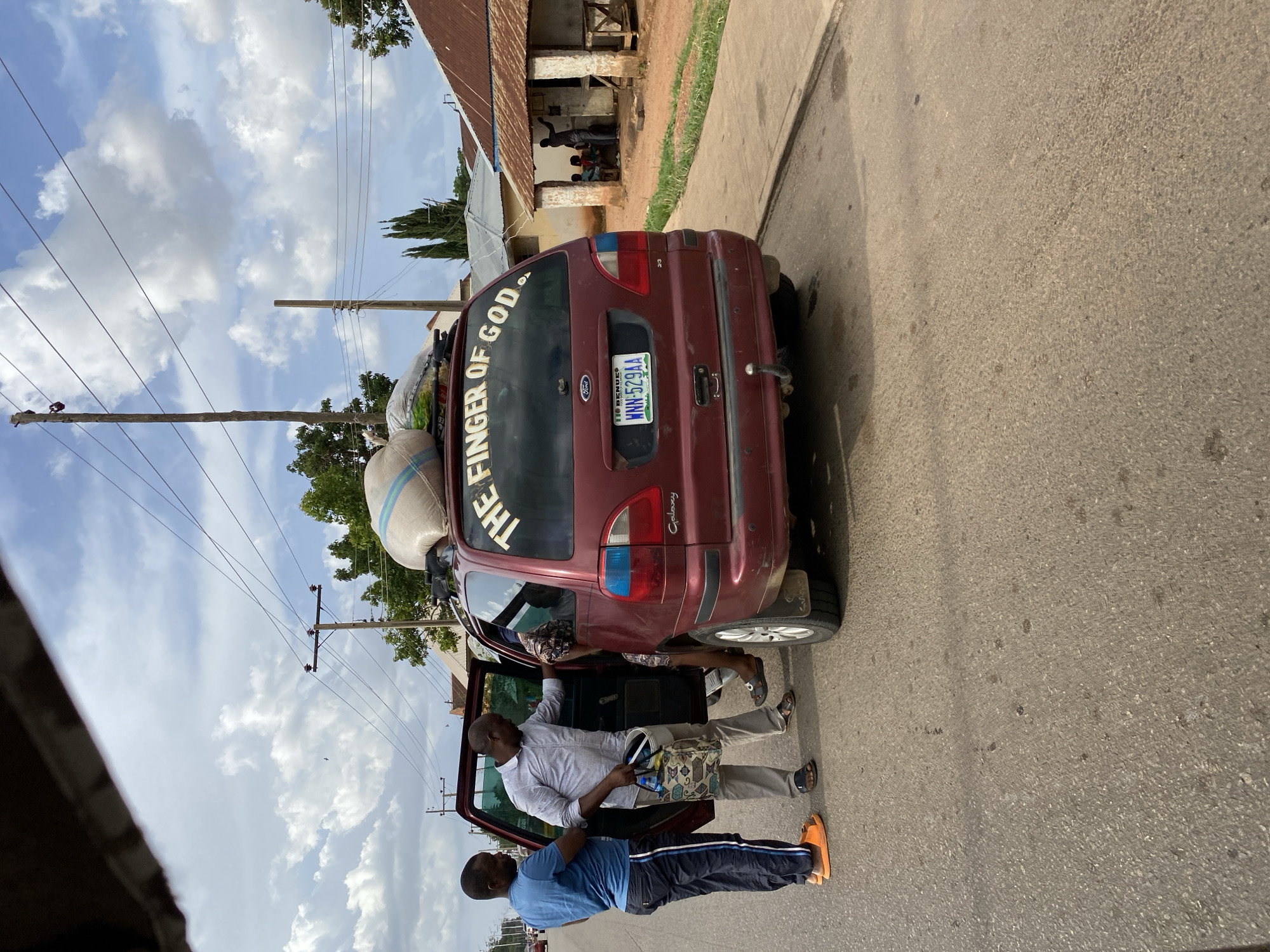 The image shows a maroon Ford Galaxy minivan parked on the side of a paved road in Bukuru, Nigeria. The vehicle has two large sacks strapped to the roof. The rear window of the minivan has a sticker that reads "THE FINGER OF GOD." A man in a light-colored long-sleeved shirt and khaki pants is standing near the open passenger door, holding a patterned bag. Another man in a blue t-shirt and dark athletic pants with orange sandals stands nearby. In the background, there is a building with a corrugated metal roof and people sitting on the porch. Power lines are visible overhead. The license plate of the minivan reads "WNN-529AA". The image is rotated 90 degrees counterclockwise.