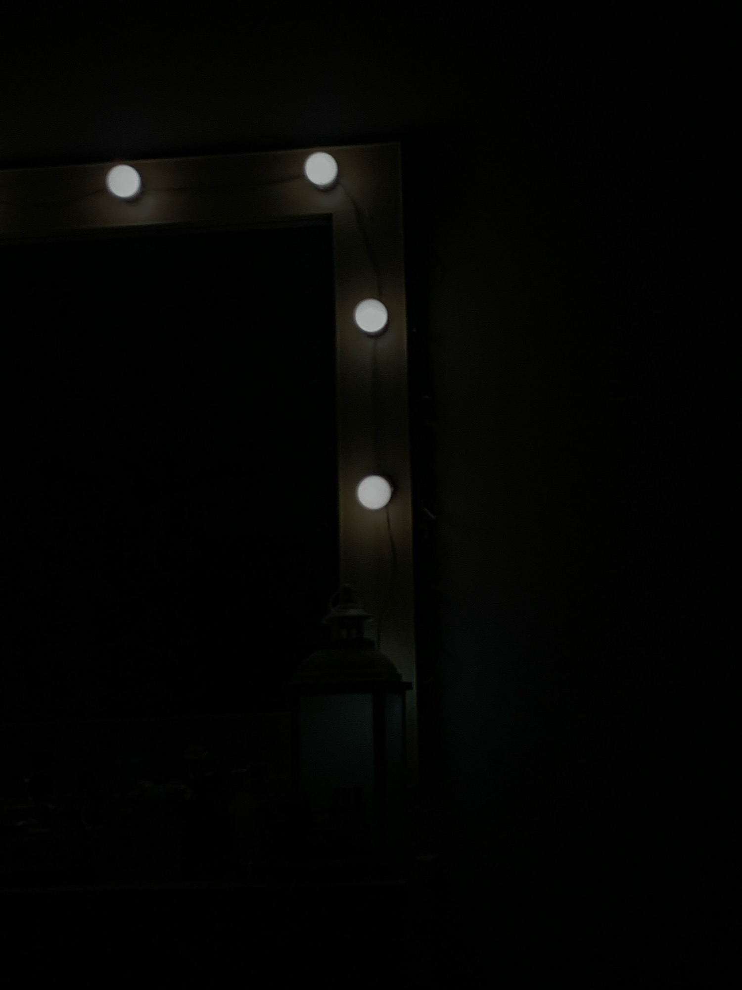 The image depicts a dimly lit vanity mirror with round lights embedded around its frame. The mirror is partially visible, reflecting darkness. A small, ornate lantern is placed on a surface below the mirror, to the right side. The background is entirely black, suggesting a dark room. There is no visible indication of the city of Islamabad, Pakistan.