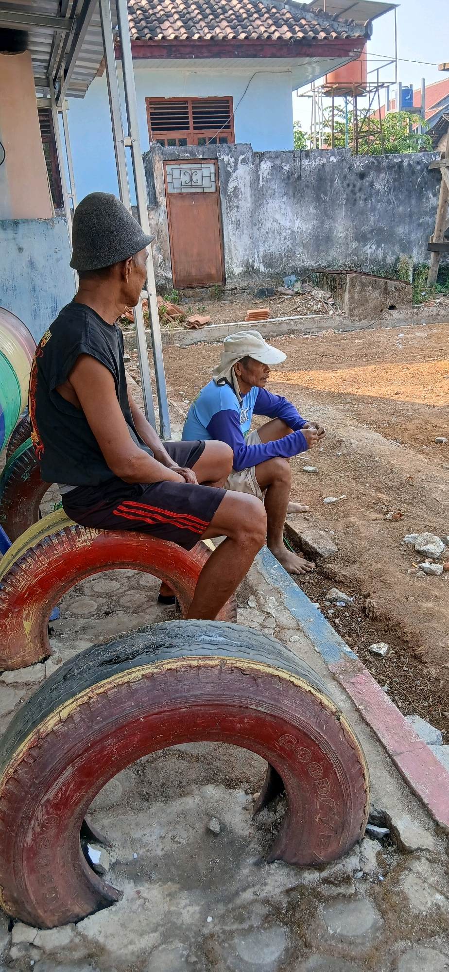 Two men are sitting on repurposed tires in an outdoor area in the city of Metro, Indonesia. The man on the left is wearing a black sleeveless shirt, black shorts with red stripes, and a grey hat. He is looking to his right. The man on the right is wearing a blue long-sleeved shirt, beige shorts, and a light-colored hat. He is looking to his right and holding something in his hands. The background shows a weathered building with a tiled roof and a water tower. The ground is dirt and gravel, with some debris scattered around. The tires appear to be old and are painted in various colors, including red and yellow.