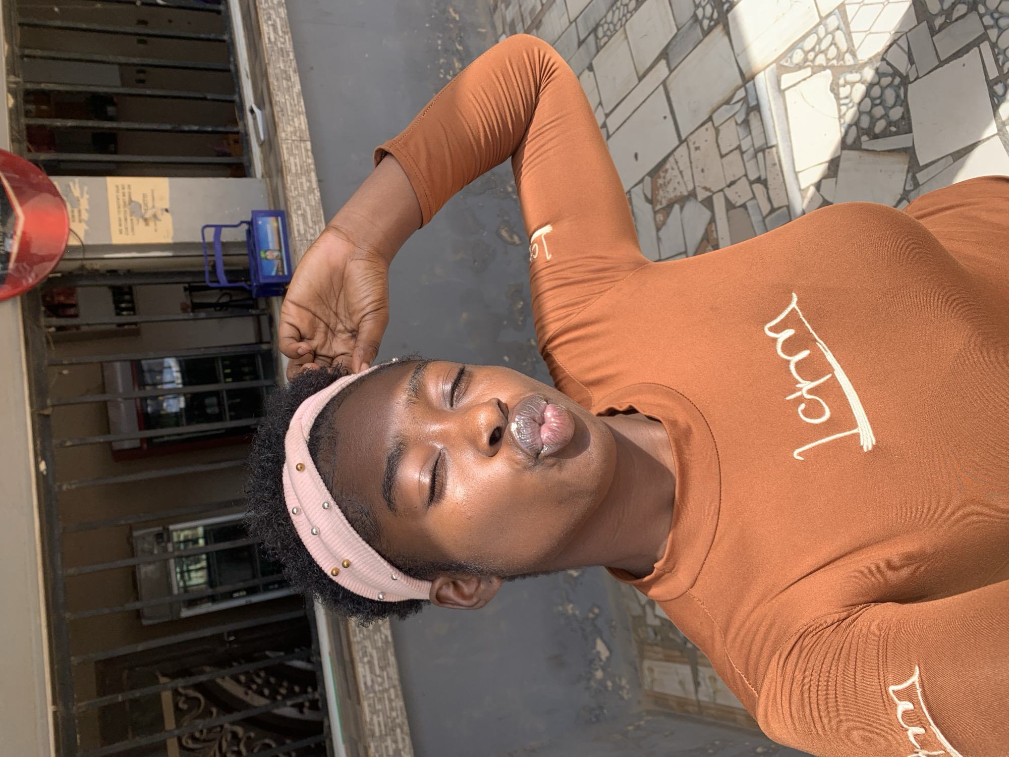 The image features a dark-skinned woman wearing a brown long-sleeved top with white writing. She is wearing a pink headband with silver and gold studs and has her eyes closed while pursing her lips as though kissing. Her right arm is raised above her head.
The background shows a building with barred windows and a red circular object mounted on the exterior. The ground is paved with textured tiles.
Based on metadata, the image location is New Stadium, Nigeria.