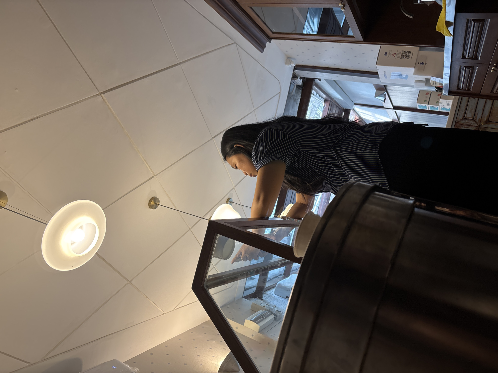 In South Jakarta, Indonesia, a woman with long, dark hair is seen from a low angle, looking up towards a glass display case. She is wearing a striped short-sleeved shirt and a dark apron. Her hands are engaged with something inside the case. The ceiling above is a white drop ceiling with integrated lighting fixtures. To the right, a wooden cabinet with glass doors is visible, containing various items, including a faucet and a yellow cloth. The environment appears to be a retail or food service establishment, possibly a bakery or deli, given the display case and the woman's attire. The background shows a window or doorway leading to another area, with more cabinets and boxes stacked on a counter. The lighting in the scene is bright, with the ceiling lights casting a warm glow.