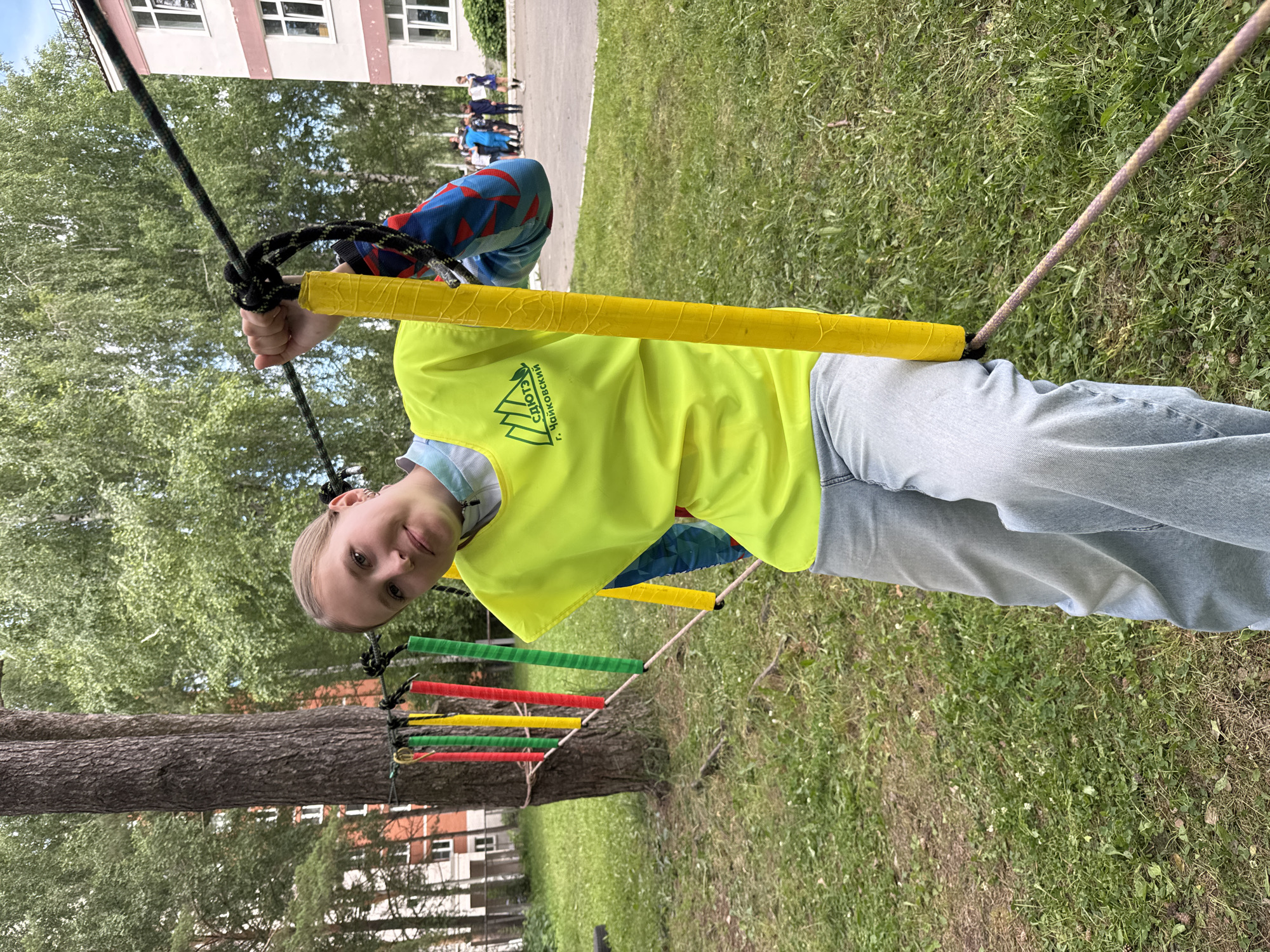 A young woman is pictured outdoors, wearing a bright yellow vest over a blue and red patterned long-sleeved shirt and light blue jeans. She is holding onto a yellow-wrapped rope handle as part of an obstacle course. Behind her, a tree with colorful fabric strips (red, yellow, green) tied to it is visible, and a rope obstacle course extends across the frame. In the background, there are buildings, trees, and other people, suggesting a public park or recreational area in the city of Chaikovsky, Russia. The ground is covered in green grass with some dirt patches. The overall lighting suggests it is daytime.