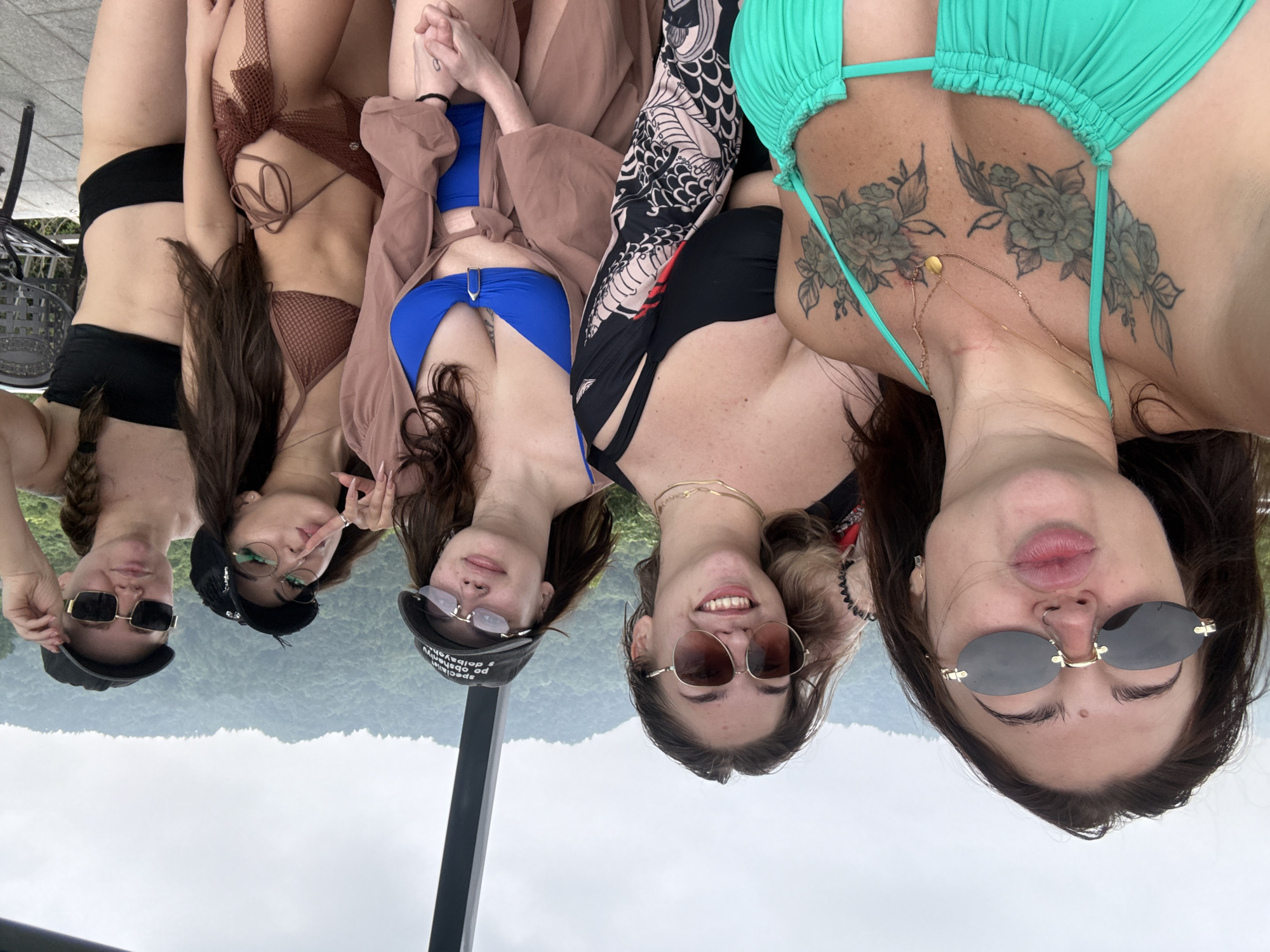 Five young women, likely in their early to mid-twenties, are posing for a selfie outdoors. The woman in the foreground is wearing a teal bikini top and large oval sunglasses, puckering her lips. To her right, a woman in a black bikini top and large round sunglasses smiles at the camera. Next to her, another woman in a black baseball cap and similar sunglasses also looks at the camera. The fourth woman, further back and to the right, wears a black baseball cap and square sunglasses. The last woman on the far right is wearing a black bikini top and has her hair in a braid.
The background shows a lush, green mountainous landscape under an overcast sky. The women appear to be on a platform or balcony, with a dark, angled structure visible on the left side of the frame, suggesting they might be in a cable car or an elevated viewing area. The presence of a metal chair and patterned floor tiles indicate an outdoor seating area or walkway.
The image does not contain any specific visual cues that would allow for precise identification of the location within Yiwu, China.