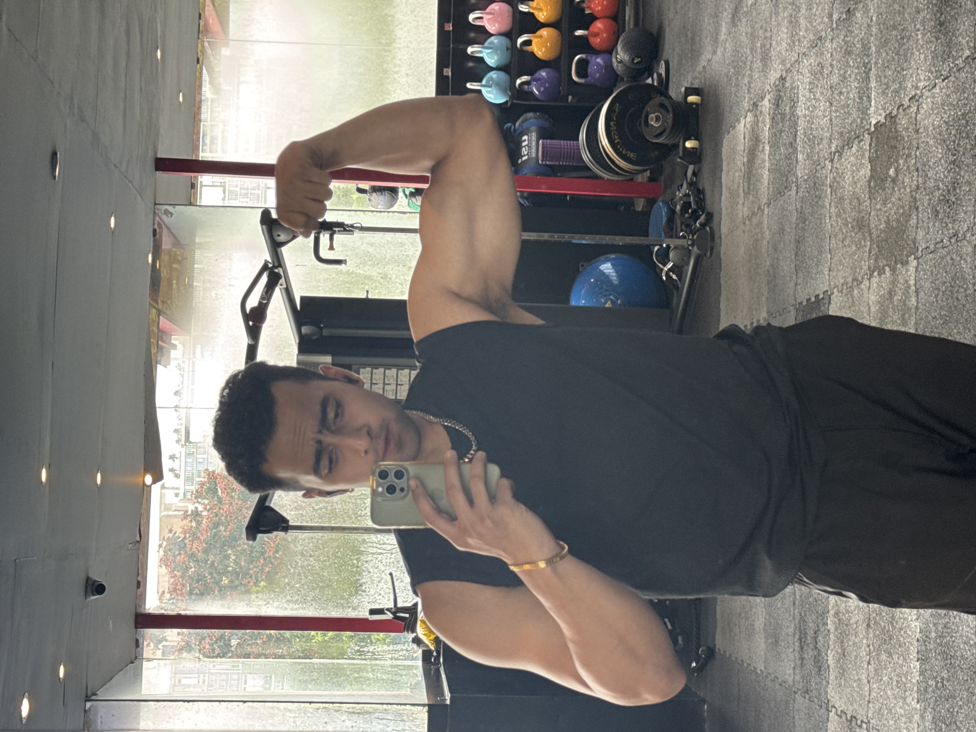 A man is taking a selfie in a gym. He is wearing a black sleeveless t-shirt and black shorts. He is flexing his right arm and looking at his reflection in the mirror. The gym appears to be well-equipped with various weight machines and free weights. There is a large window in the background, showing a glimpse of an urban environment with trees and buildings. The floor is covered with grey interlocking rubber mats. The lighting in the gym is a combination of overhead spotlights and natural light coming from the window. The overall ambiance suggests a typical fitness facility. Based on the visible architecture and vegetation in the background, it is plausible that this gym is located in Kolkata, India, given the tropical-like trees and the style of buildings visible.