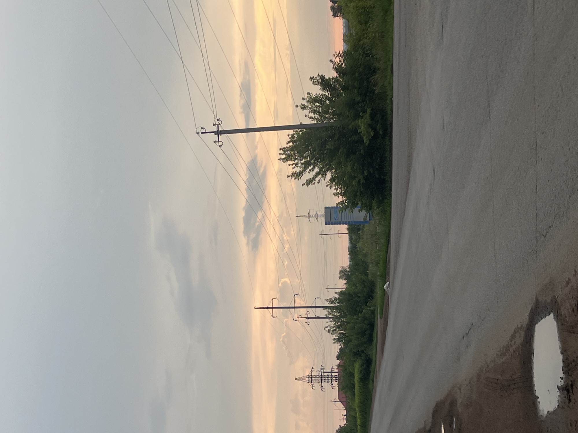 The image captures a roadside scene with power lines stretching across a cloudy sky. The sky is illuminated by the soft, warm hues of either sunrise or sunset, with streaks of orange and pink visible through the pale grey clouds. The scene is dominated by a paved road running diagonally from the lower right to the upper left corner of the frame.
To the left of the road, a line of trees and lush green vegetation forms a dense border. Several large electricity pylons with numerous wires are interspersed along this line, marching into the distance. One pylon is particularly prominent in the mid-ground, with its crossbars supporting the overhead cables. Further down the road, another, more elaborate lattice-style pylon is visible, and a blue and white billboard can be spotted behind the trees.
The road surface appears wet in places, with a large, reflective puddle prominent in the lower right corner, suggesting recent rain. The overall mood is tranquil and slightly atmospheric, perhaps indicating the end of a day or the beginning of a new one. No people are visible in the frame, but the presence of the road, power lines, and billboard suggests a human-modified landscape, likely on the outskirts of a town or rural area. The location context of Buzuluk, Russia, provides a geographical anchor for this scene.