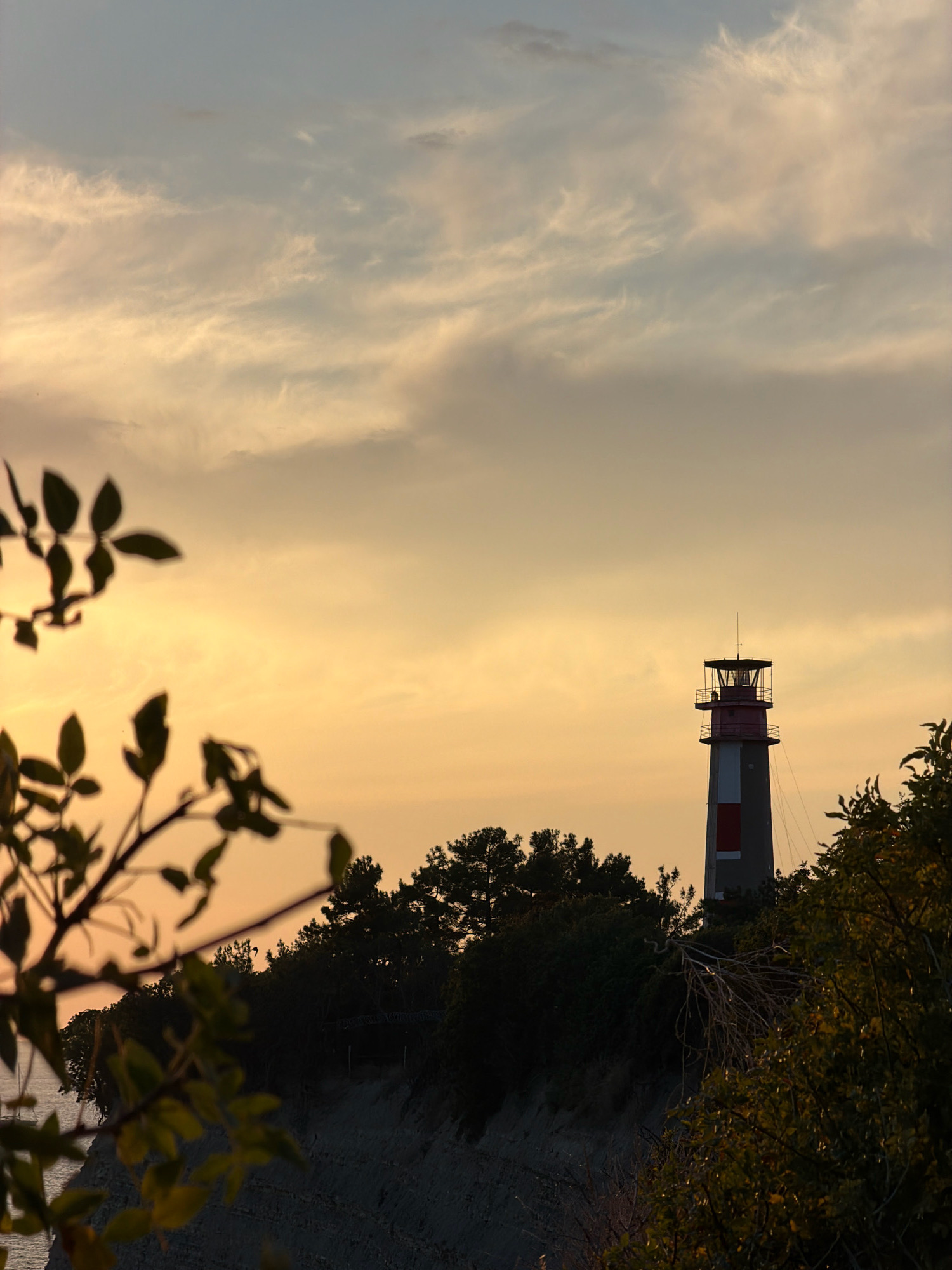 This is a photograph of a lighthouse on a clifftop overlooking the sea during a sunset. The sky is filled with soft, pastel clouds in shades of orange, yellow, and pale blue. In the foreground, the silhouette of a leafy branch is visible on the left, partially obscuring the view. The lighthouse is tall and cylindrical, with red and white bands and a dark top. It is surrounded by dark green trees and vegetation. The ground appears to be a rocky cliff face. The overall atmosphere is serene and picturesque. This lighthouse is located in Gelendzhik, Russia, specifically on Tolstoy Cape, which is a well-known landmark in the city. There are no people visible in the image.