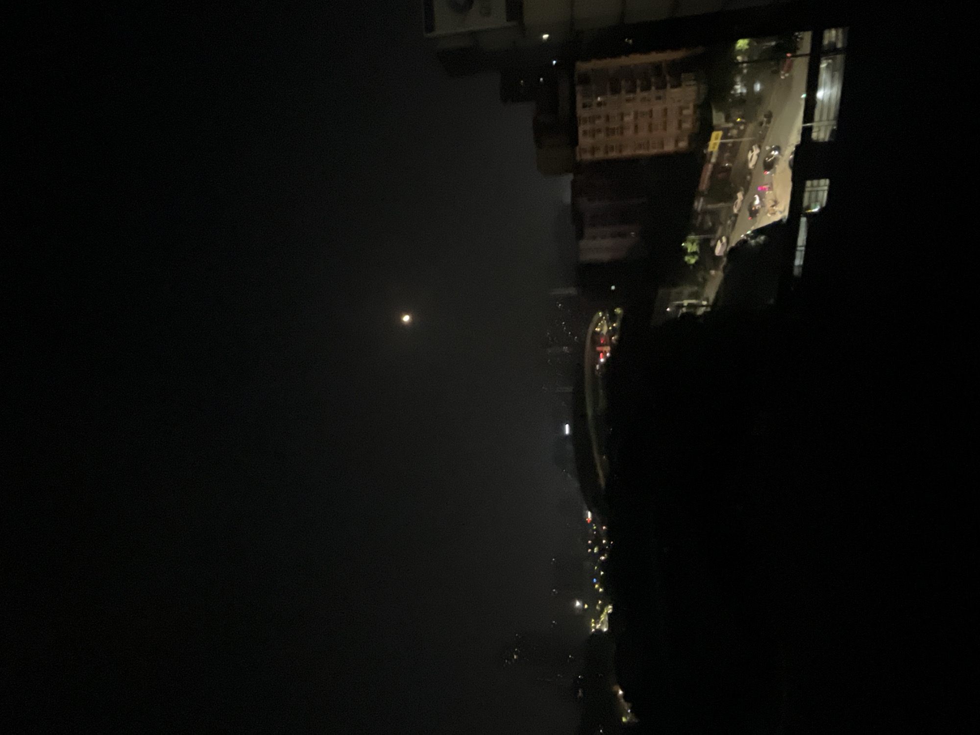 The image displays an elevated nighttime view of an urban area within Jiulongpo District, China. The dark sky features a single, bright, circular moon in the upper center. Below, a vast cityscape stretches into the distance, characterized by numerous scattered artificial lights and a faint atmospheric glow. In the mid-ground, a large, dark, domed structure is visible, consistent with a stadium or major building.
On the right side of the frame, several multi-story buildings are prominent, including a lighter-toned building with multiple visible windows and a reddish-brown building further left. A large, partially visible sign is present on the far right edge. A street or road occupies the lower mid-ground, running diagonally across the frame. Multiple vehicles are visible on this street, including a white van, several white sedans, and a black sedan. A distinct bright pink/magenta element, appearing to be a person in bright clothing or a reflective object, is centrally located on the street among the vehicles. Faint human figures are also discernible on the sidewalk or near the street. The immediate foreground on the left and bottom is largely dark and indistinct.