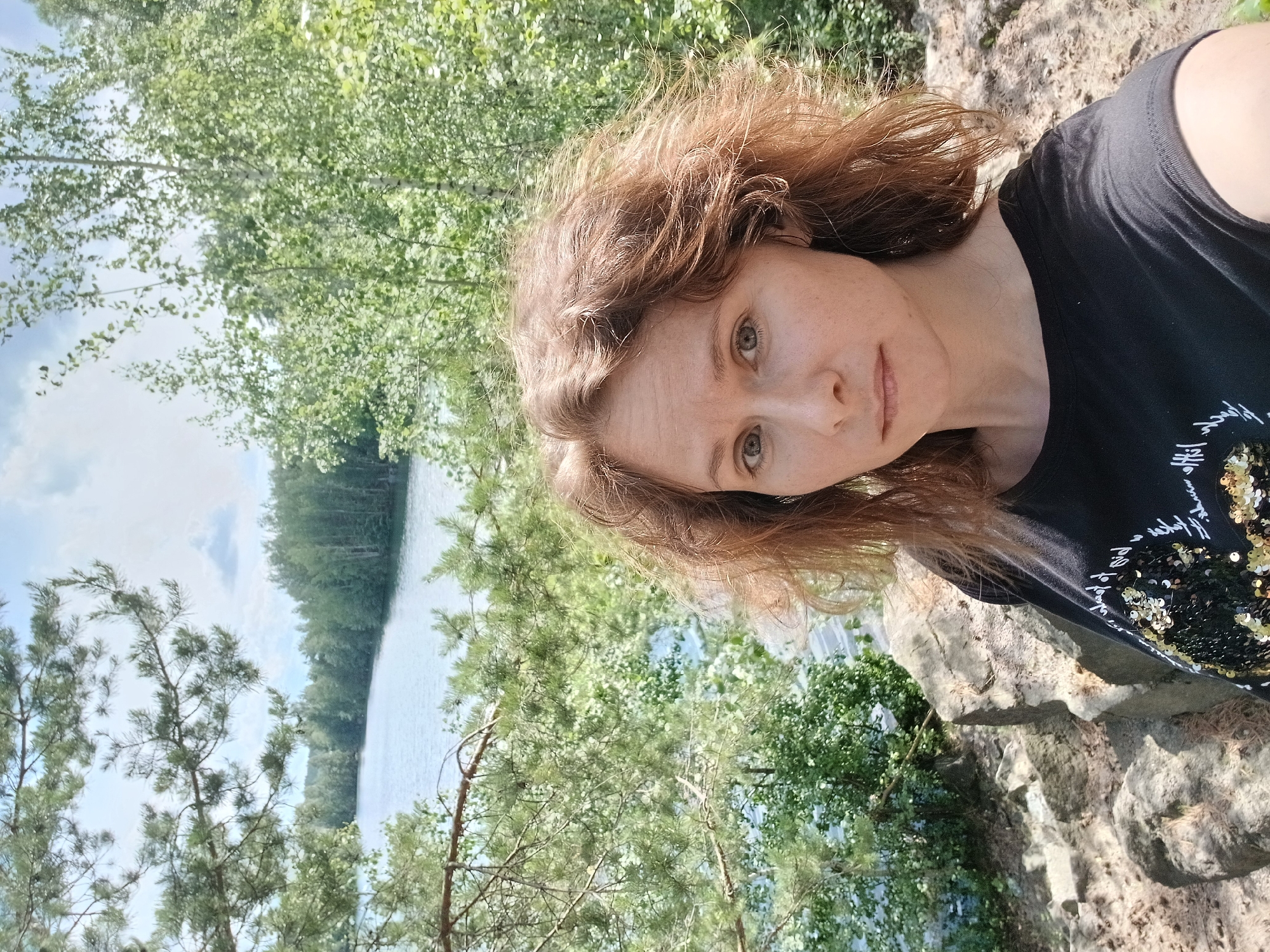 A woman with reddish-brown hair is taking a selfie outdoors by a lake. She is wearing a black t-shirt adorned with gold and black sequins in a circular pattern. The background features a body of water, lush green trees, and rocky terrain. The sky is visible with some clouds. The location is the city of Yekaterinburg, Russia.