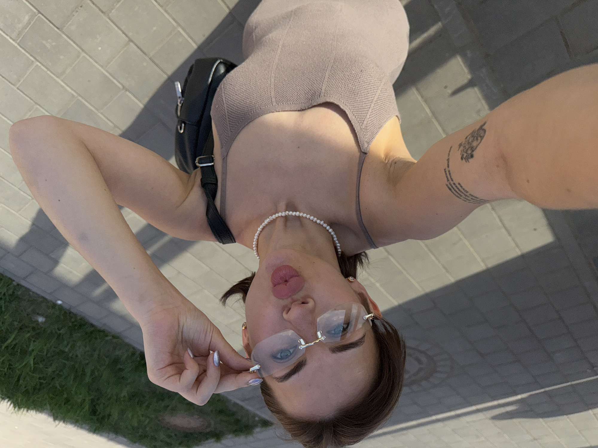 A woman stands on a paved surface in Sarapul, Russia, taking a selfie. She wears a sleeveless, fitted dress, a pearl necklace, and rimless glasses. A black backpack is slung over her shoulder. The woman has a tattoo on her right arm and manicured fingernails. She puckers her lips and holds her glasses with her left hand. Grass is visible in the lower left corner of the frame. The pavement is likely part of a sidewalk or pedestrian area in the city.
