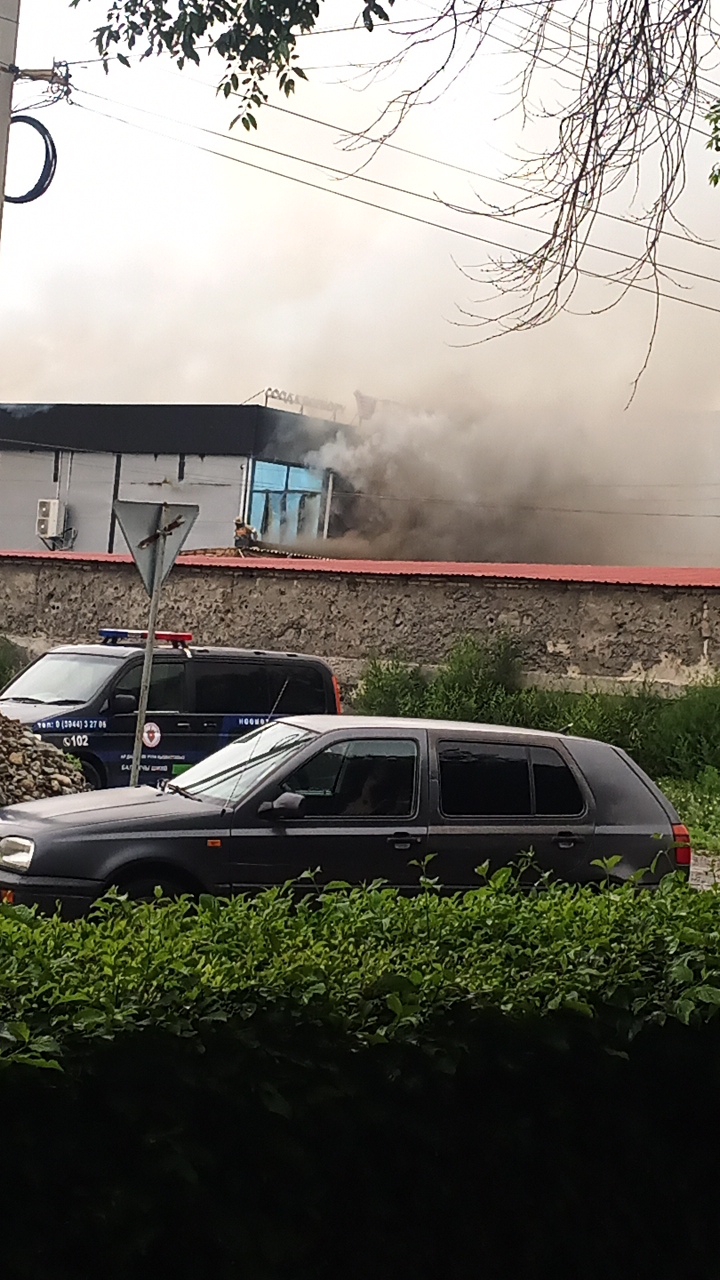 In Balykchy, Kyrgyzstan, on a cloudy day, a building is engulfed in thick, dark smoke emanating from its windows. The building appears to be a commercial or industrial structure with a red roofline and grey walls. A police car, distinguished by its blue and red lights, is parked in front of the building. In the foreground, a dark-colored hatchback car is visible, partially obscured by lush green foliage. The overall scene suggests an active emergency situation, likely a fire, with emergency services present.
