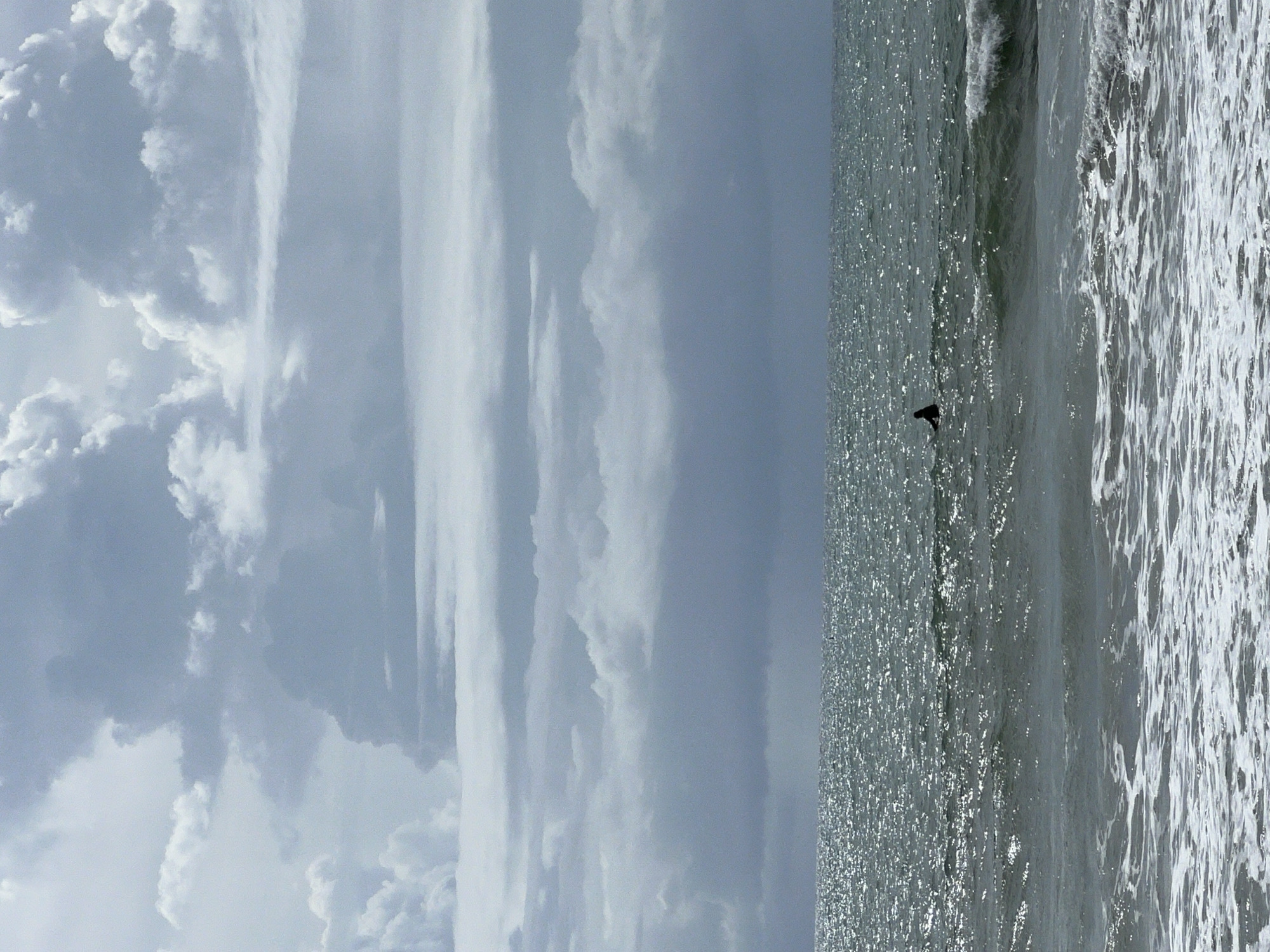 The image displays a wide view of an ocean under an overcast sky. The upper two-thirds of the frame are filled with various formations of grey and white clouds, some appearing denser and darker than others. The horizon line is clearly defined and flat, separating the sky from the ocean. The ocean surface is dark grey-green, with numerous small specular highlights indicating reflected light, creating a glittering effect. In the mid-ground, slightly to the left of the frame's center, a single person is visible, silhouetted in dark clothing, sitting upright on a surfboard. Small, breaking waves with white foam are present in the foreground, with the water surface becoming choppier further out. The scene takes place in the city of Unknown, Unknown.