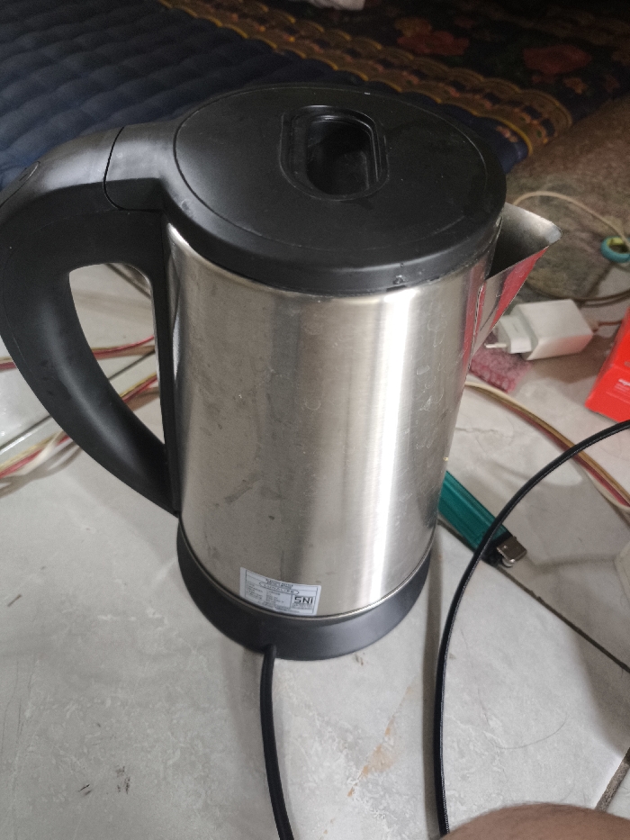 A stainless steel electric kettle with a black handle, lid, and base sits on a tiled surface. The kettle has a spout and a small label near the base. Various objects are scattered around the kettle, including cables, a lighter, a phone charger, and what looks like part of a phone package. A bed or couch with a patterned cover is visible in the background.