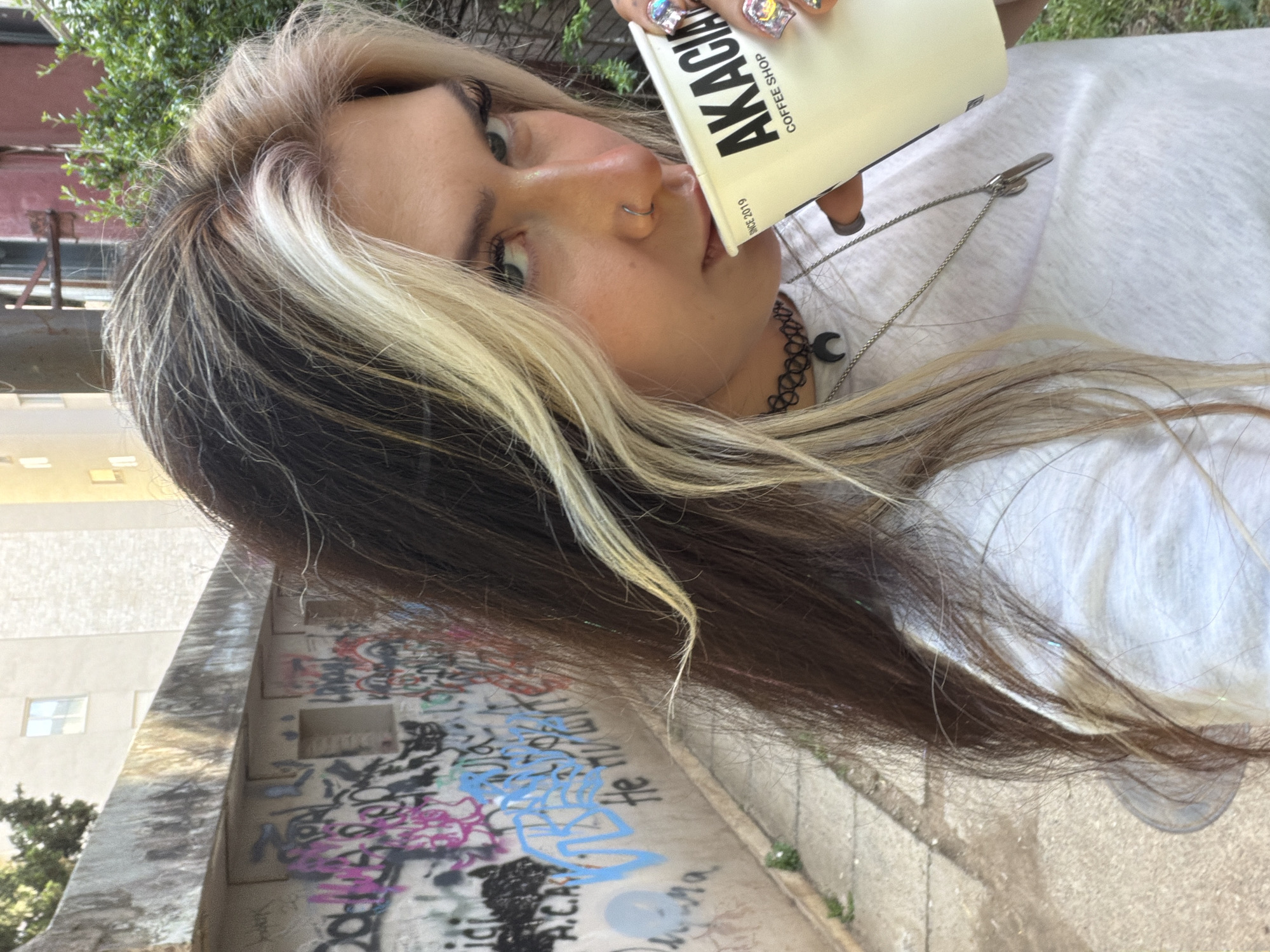 A brightly lit, slightly tilted close-up portrait captures a young woman outdoors in Budva, Montenegro. She is looking directly at the camera while holding a light-colored coffee cup to her mouth.
The woman has long, straight hair featuring prominent blonde streaks that frame her face, contrasting with her darker brown hair. She wears a nose ring and dark eye makeup. Her fingernails are notably long and elaborately decorated with numerous iridescent gems and embellishments. She is dressed in a light-colored, possibly white, top and accessorizes with two necklaces: a black choker adorned with a small crescent moon charm, and a longer, thin silver chain with a simple, elongated pendant.
The coffee cup she holds clearly displays the text "AKACIA COFFEE SHOP" in bold black capital letters, with "SINCE 2019" printed below.
The background features a concrete or stone wall covered extensively in colorful graffiti, showcasing various tags, letters, and symbols in shades of blue, pink, and black. A partially legible phrase in blue appears to read "He HUXLONT." Beyond the graffiti-covered wall, sections of a building with windows are visible, and hints of green foliage suggest the presence of trees or bushes in the outdoor setting. The ground in the foreground is paved, contributing to the urban environment. The image has a candid feel, captured during daytime under bright conditions.