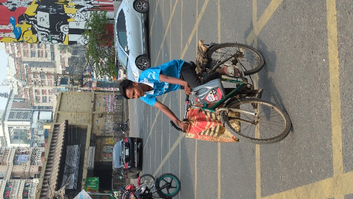 A person on a bicycle in the street, holding a bottle of oil. The bicycle has a red and gold cloth hanging over the front. Behind him are cars and motorcycles parked on the street. In the background are buildings and a mural. The street is marked with yellow lines. The person is wearing a blue shirt and black pants. He is wearing sandals.