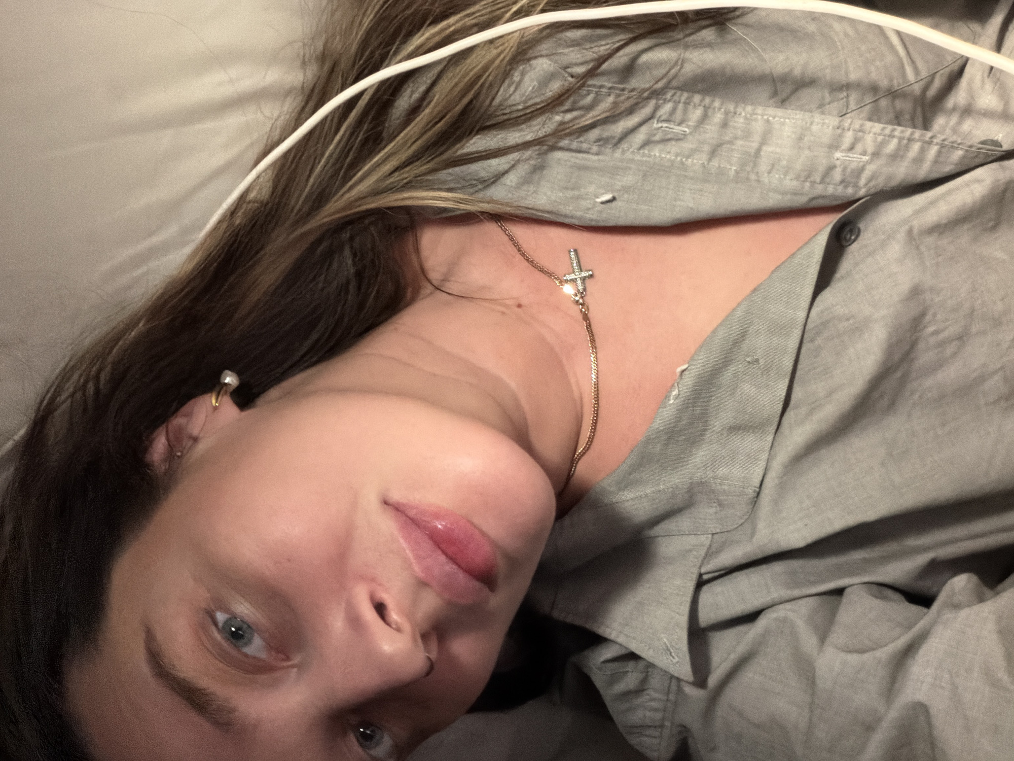 A person is lying down wearing a grey collared shirt with the top buttons undone, revealing a gold necklace with a cross pendant. They have light skin, blue eyes, and brown hair with blonde highlights. A white wire is draped over their head. The background consists of grey fabric, possibly bedding. There is no indication of the location being Moscow, Russia.