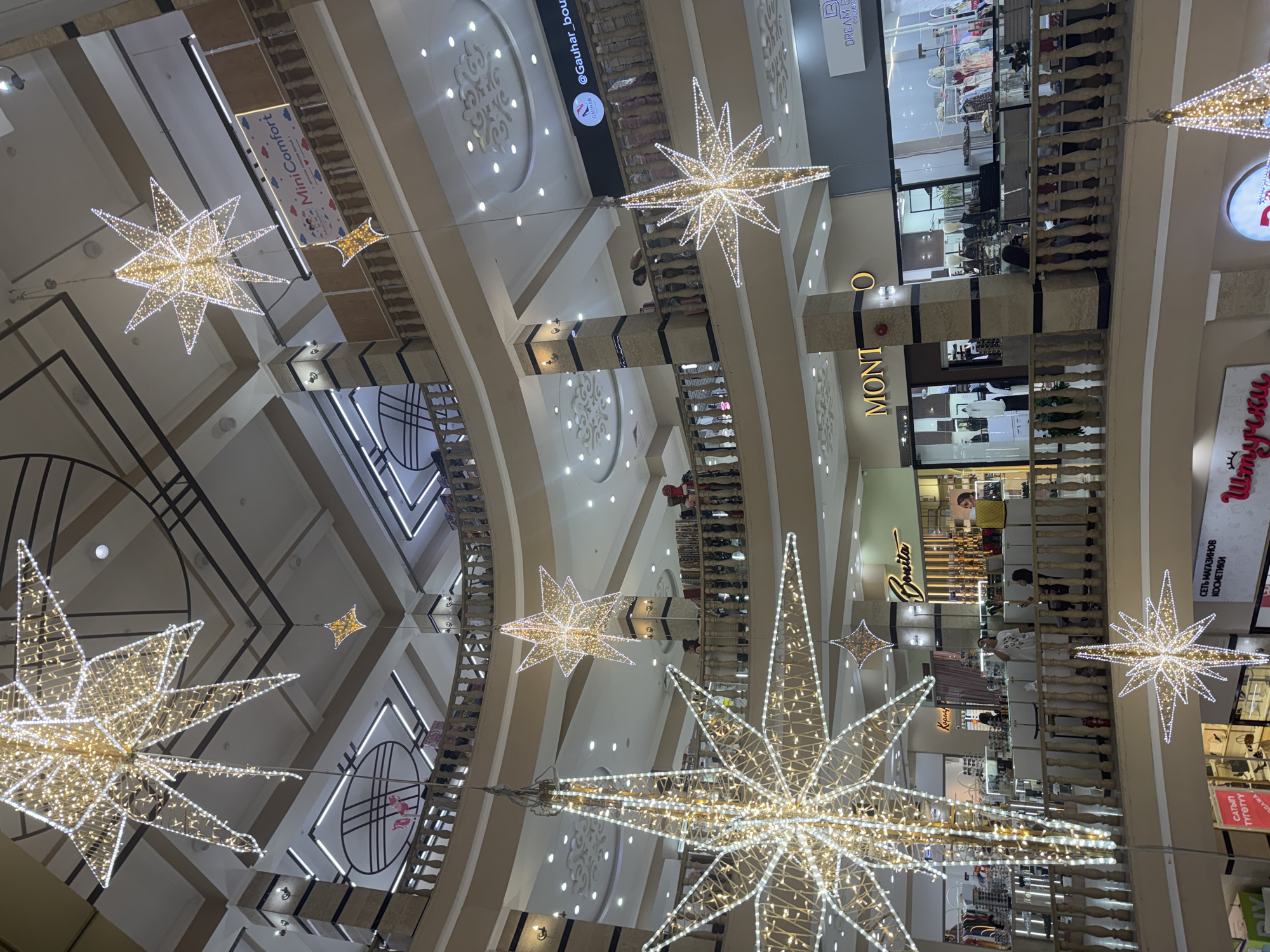 The image shows the interior of a multi-level shopping mall in Bishkek, Kyrgyzstan, decorated with large, illuminated star-shaped ornaments. The mall features a light-colored, ornate interior with balconies on multiple levels. Various shops are visible on the different floors, including signs for "Bonita," "MONTO," and "Dream Beauty Collection." People are seen browsing in the shops and walking along the balconies. The lighting suggests it is daytime or the mall is well-lit. The overall impression is of a modern, upscale shopping environment adorned for a festive occasion.