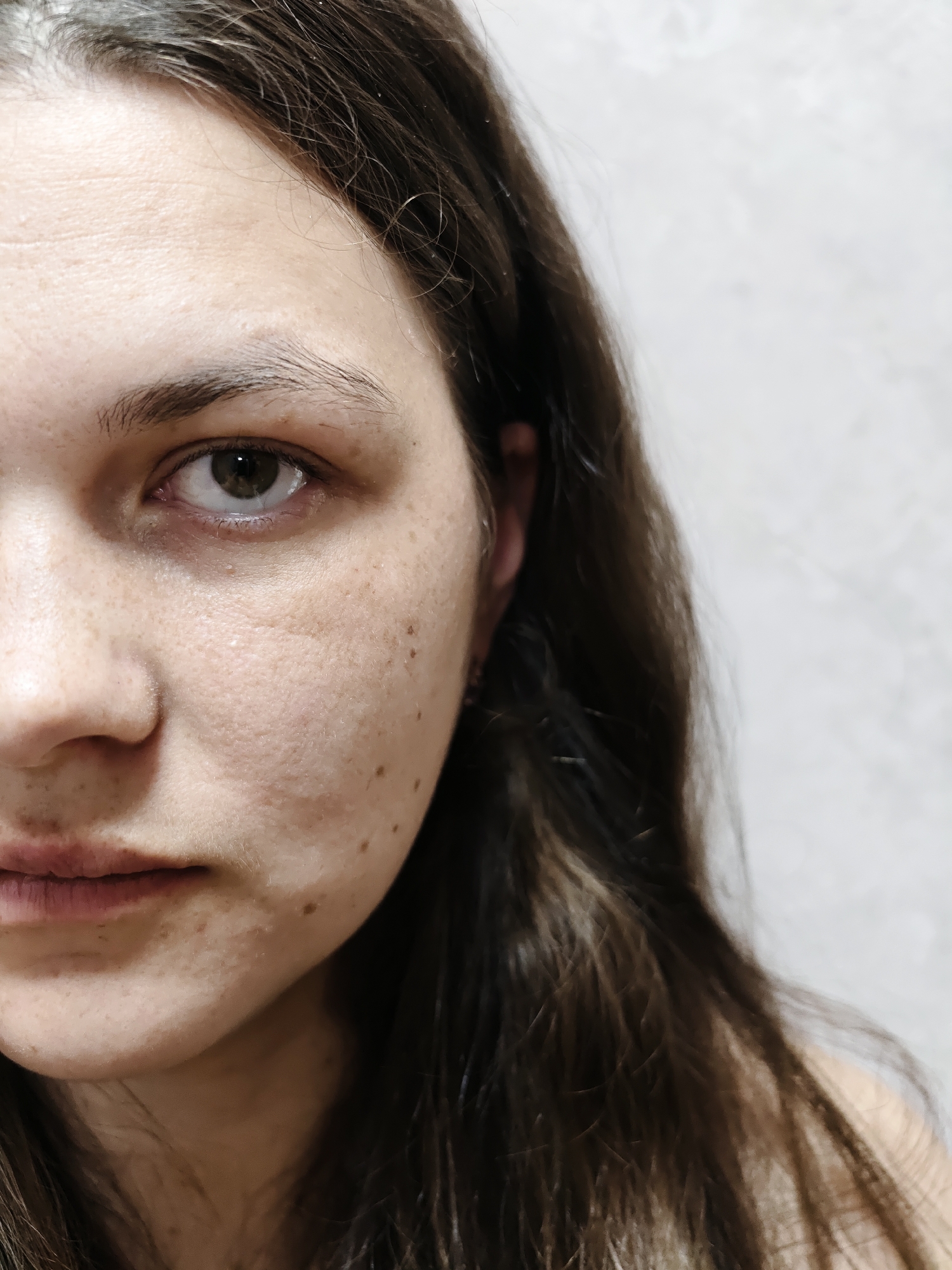 A close-up, half-portrait photograph features the left side of a person's face, likely a woman, looking directly into the camera. The subject has fair skin, dark brown hair, and a neutral expression. Only her left eye is fully visible, displaying a dark green or hazel iris with a defined pupil. Her dark eyebrow is naturally shaped.
Notable details of her skin include visible pores across her cheek, nose, and chin, along with several scattered dark brown freckles or moles. There are also a few small, light-colored bumps visible near the lower lash line of her left eye and on her cheek. Fine, light-colored facial hair is discernible on her upper lip and chin area. Her lips are a natural reddish-pink hue, with only the left side of her mouth visible. Her dark brown hair cascades down the right side of the frame, partially obscuring her ear and shoulder, and appears to be long and somewhat wavy or curly. The background is a plain, light-colored wall with a subtle texture, suggesting an indoor setting in Chaikovsky, Russia. There is no visible text in the image.