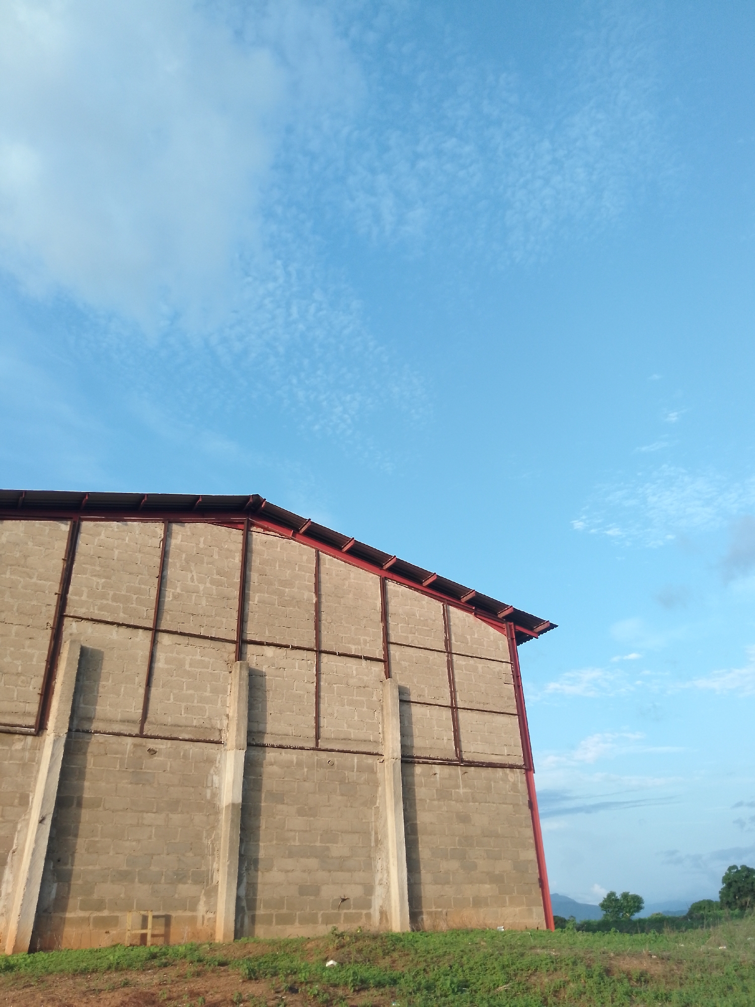 The image depicts the side of a partially constructed building in Serti, Nigeria. The building features a cinder block wall with a red metal frame supporting a corrugated metal roof. Vertical concrete pillars are visible along the exterior of the wall, casting shadows. The ground in front of the building is covered with green grass and some sparse vegetation. In the background, a light blue sky with scattered cirrus clouds dominates the upper portion of the image. Distant hills and trees are visible on the right side, suggesting a semi-rural or developing area. There are no people or animals present in the image.