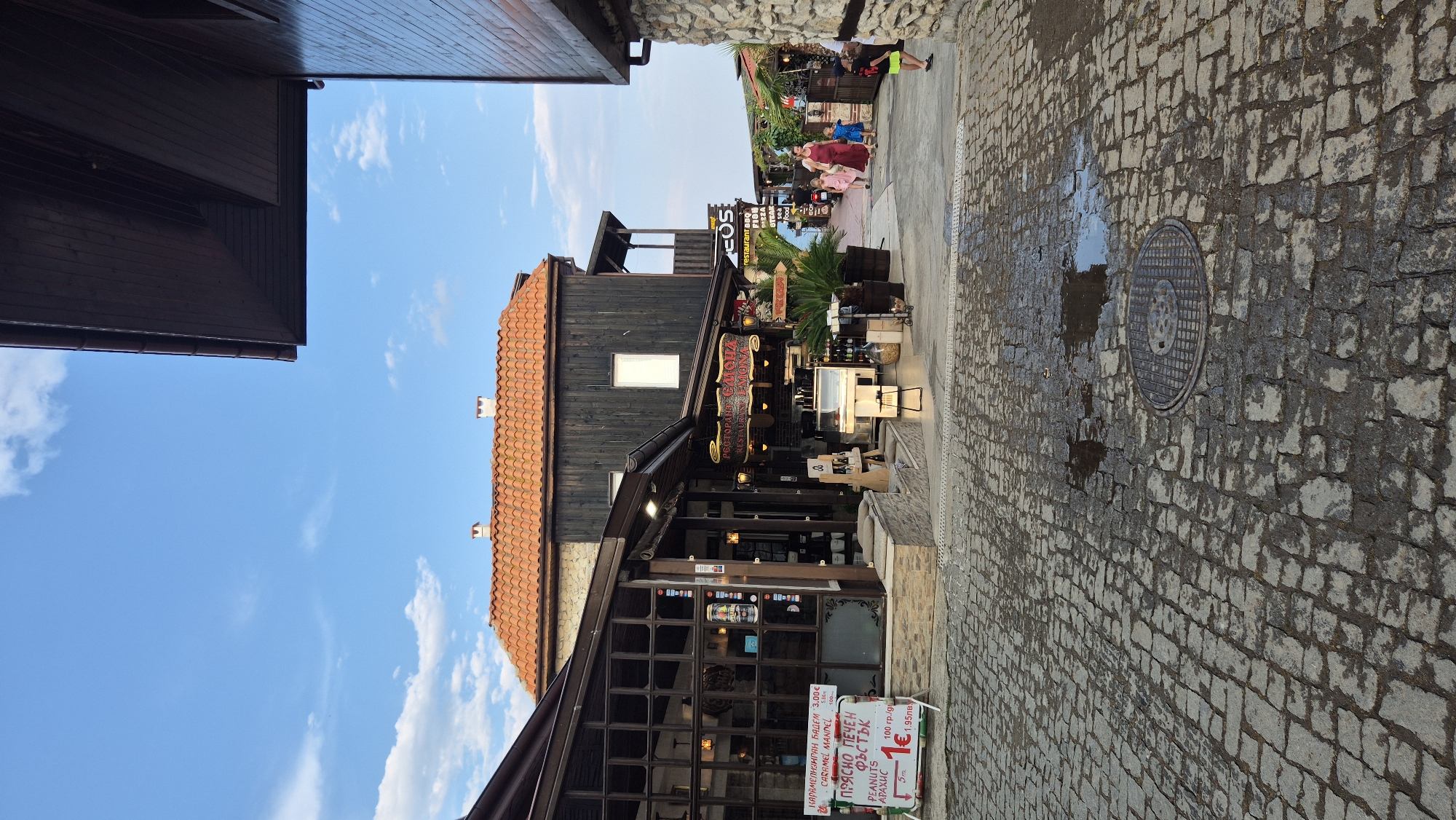 The image shows a street scene in the old town of Nesebar, Bulgaria, with cobblestone paving wet from recent rain. The buildings are traditional, with dark wooden facades and red tile roofs. One building on the left has large glass windows, likely housing a shop or restaurant. A sign in Cyrillic and English displays prices for "CARAMEL MANDEL" and "PEANUTS." Further down the street, a restaurant sign reads "RESTAURANT LEMONA." A family with two young children is walking away from the camera down the street towards a cluster of shops. The sky is blue with scattered white clouds.