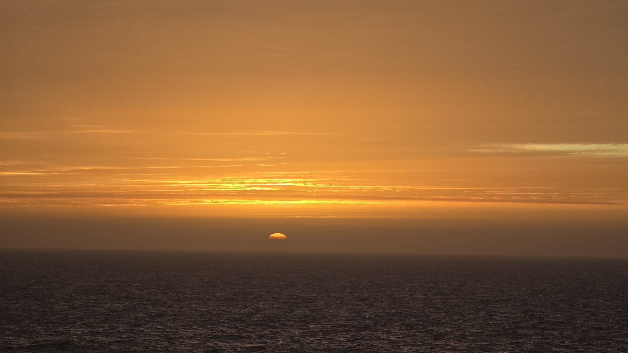 A wide shot displays a sunset over the ocean. The sky is a mix of orange and yellow hues, with some wispy clouds near the horizon. The sun is setting over the water, appearing as a semicircle. The sea is dark and stretches out into the distance. There is no discernible city or people in the image.