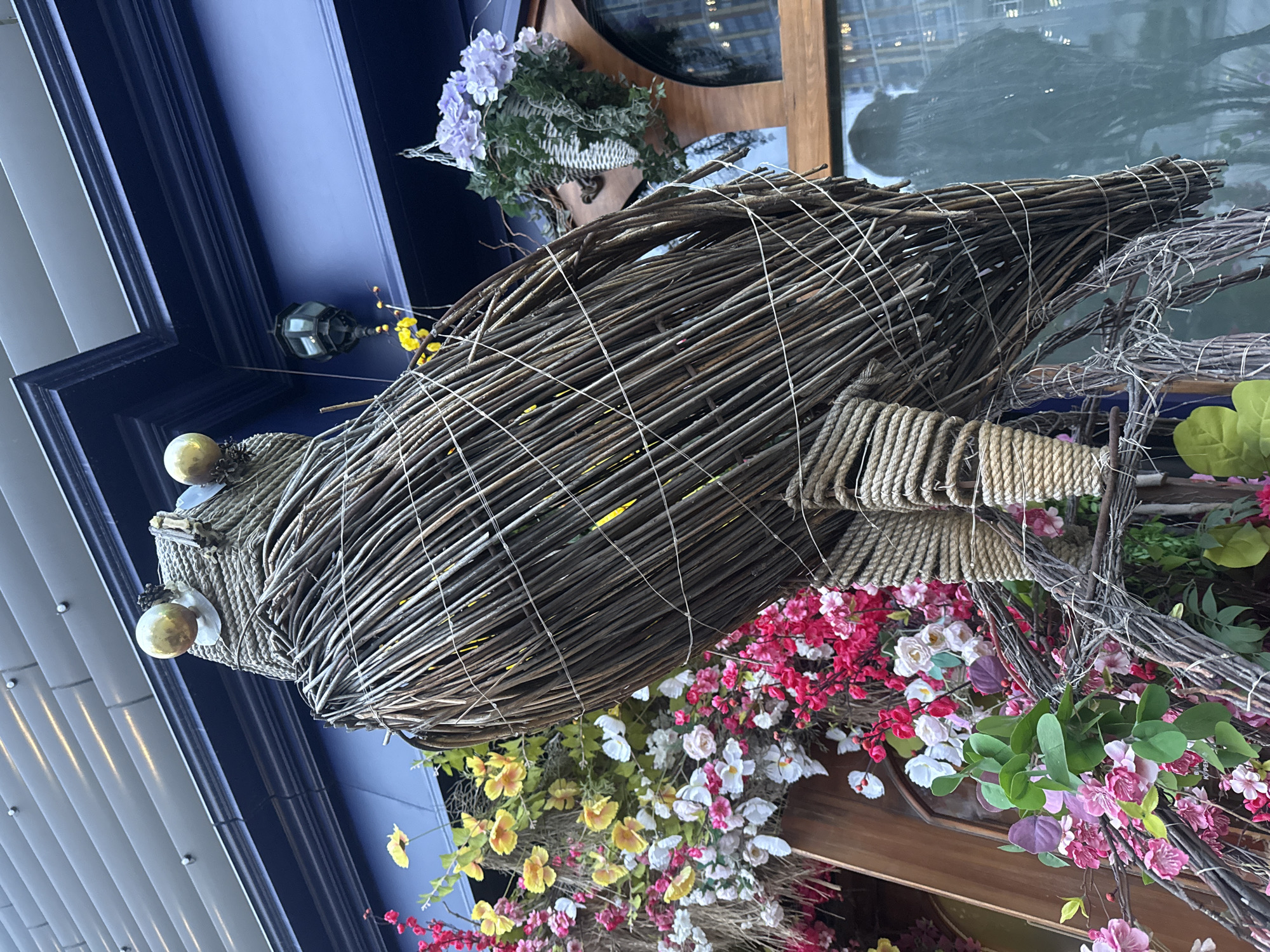 A large, decorative bird sculpture, crafted from interwoven dark branches and twigs, dominates the central and right portions of the image. The bird's body is constructed from numerous thin, dark brown sticks bound with lighter string, and sections of its form are wrapped with thick, coiled rope. Its head features two spherical, light-colored eyes, possibly golden, and a small, conical beak.
The bird is surrounded and partially integrated with a vibrant display of artificial flowers in various hues including bright pink, white, yellow, and light purple, complemented by green foliage. These floral arrangements appear to be part of the same decorative installation.
In the upper left background, a dark blue architectural element, possibly a window frame or part of a building facade, is visible. A small, dark lantern or light fixture hangs near this blue structure. To the far left, light grey building panels are present. Behind the bird and flowers, a reflective window surface shows blurry reflections of modern buildings, indicating an urban setting.
The image captures an elaborate outdoor decorative display, likely adorning a shopfront or building exterior. The lighting is soft and diffused, suggesting it might be an overcast day or indirect light. No specific activity or event is taking place; it is a static display. There is no visible text identifiable in the image. The scene depicts an aesthetic enhancement for a location in Yekaterinburg, Russia.