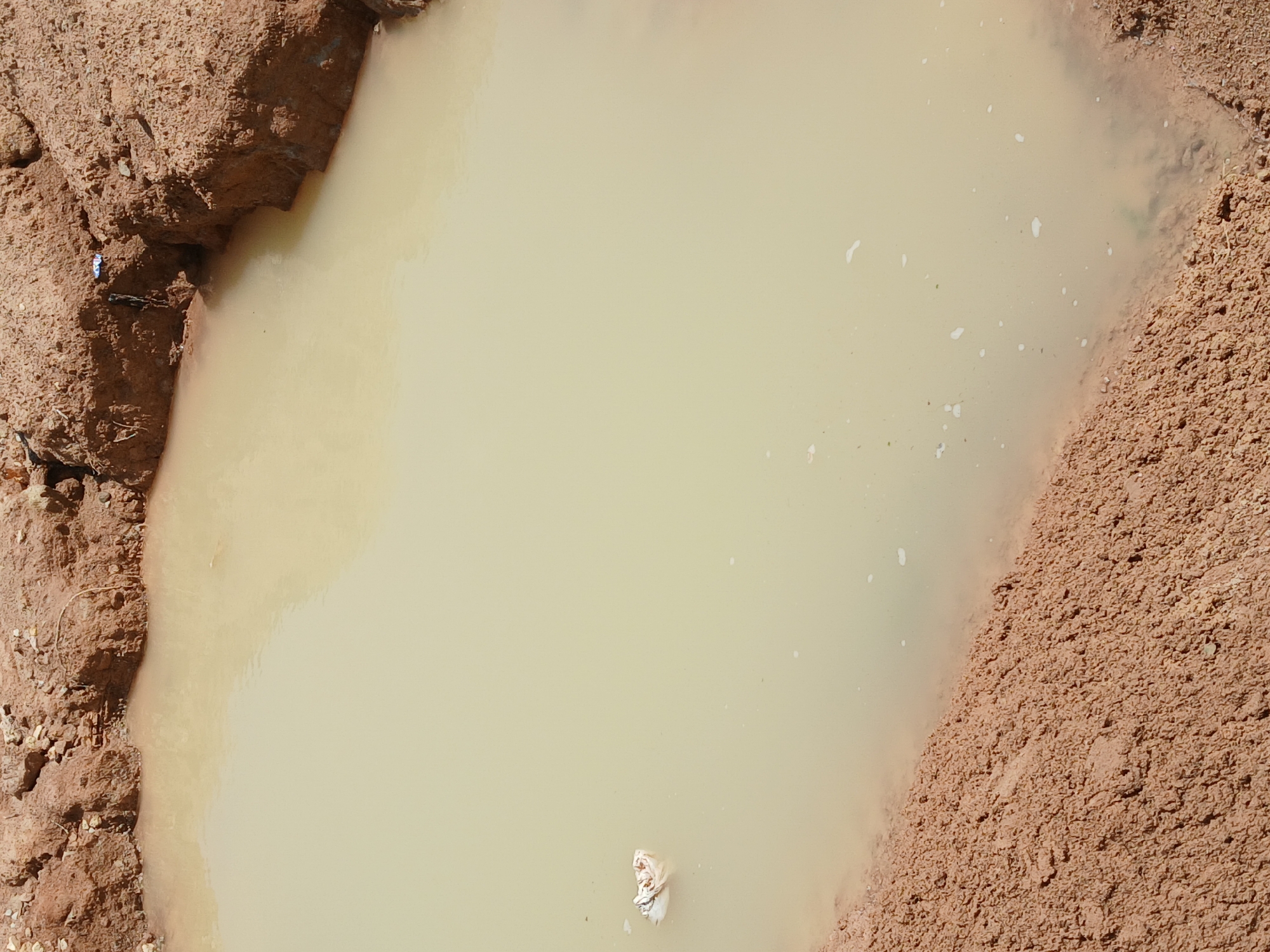 The image displays a murky, light-colored pool of water with muddy banks on the left and right sides. The water appears to be shallow and contains small white specks, possibly debris or sediment. A crumpled piece of white material is floating in the water towards the bottom center.
The left bank is rough and composed of reddish-brown soil with some small rocks and sparse vegetation. A small piece of white and blue plastic and some dark, thin objects are visible on this bank. The right bank is also made of reddish-brown soil but appears more granular and less consolidated than the left bank.
The scene suggests an outdoor environment, likely in a natural or semi-natural setting due to the presence of mud and water. The lighting indicates it is daytime, with the sun casting a bright light that creates some sheen on the water's surface. There are no people visible in the image, and the activity taking place is simply the stillness of the water and the surrounding environment. No text is visible in the image. The location context is Katon Rikkos, Nigeria.