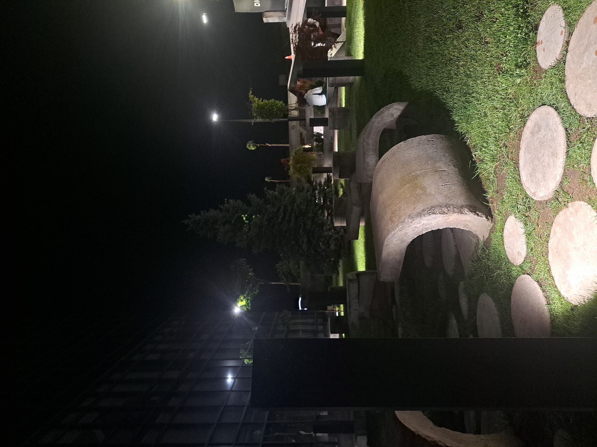 The image shows an outdoor scene at night. A large, concrete sculpture is partially visible on the right, surrounded by grass and a path of stepping stones. The sculpture appears to be arch-shaped. In the background, there are trees and streetlights. In the mid-ground, there are benches and decorative plantings, where a person is seated. In the distance, there is a building with a grid-like facade. The location is the city of Yerevan, Armenia.