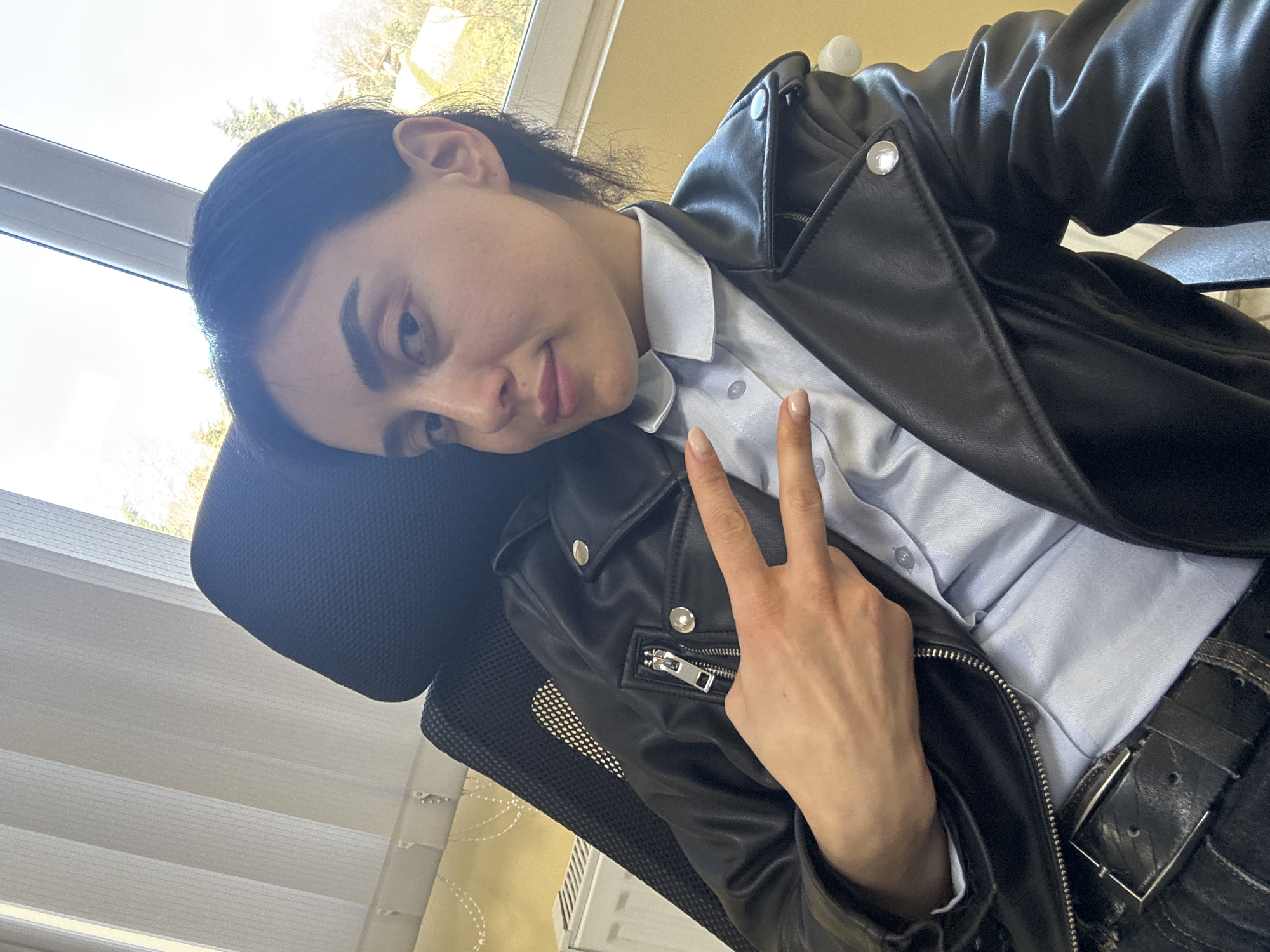 A person is seated in a black office chair, leaning their head against the headrest. They are wearing a light blue button-down shirt and a black leather jacket. The person's hand is raised, making a peace sign. The chair is positioned near a window with blinds. The location is likely indoors, with hints of greenery visible through the window. While it is not explicitly shown, based on the general features of the environment and typical interior decor, it is reasonable to infer the location may be Tbilisi, Georgia.