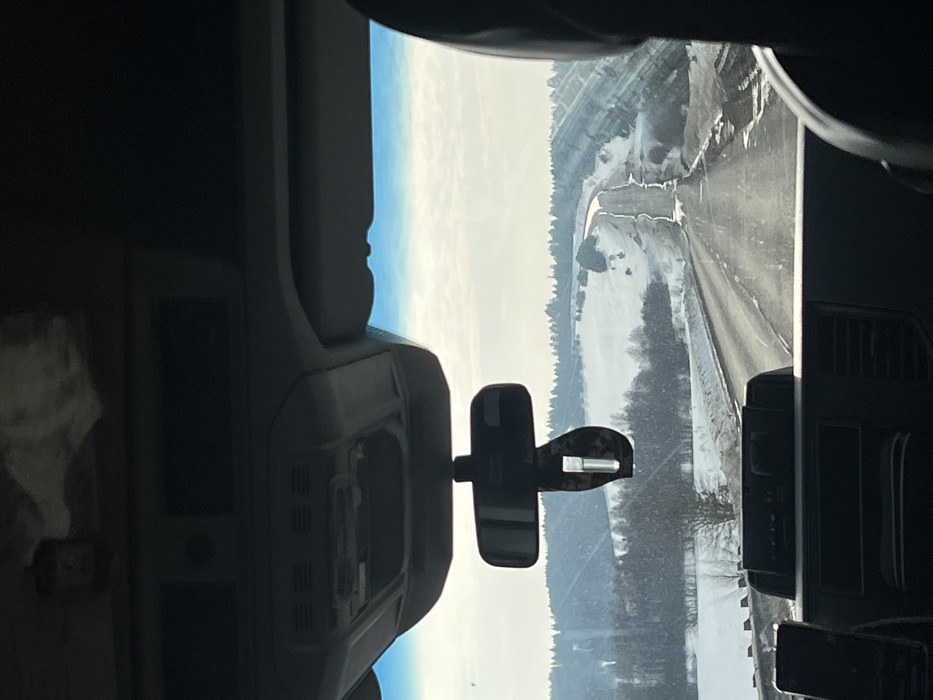 The image, captured from the interior of a vehicle looking forward through the windshield, depicts a winter landscape near Samolet, Russia. The foreground includes the vehicle's dark gray dashboard and an overhead console with various vents and compartments. A rear-view mirror is centrally positioned, from which a dark, rectangular air freshener hangs. No individuals are visible.
The scene outside features a two-lane asphalt road, partially cleared of snow, extending into the distant rolling hills. Snow and slush are present on the road's shoulders and median, and in some areas, partially cover the driving surface. The surrounding landscape consists of snow-covered fields and hills, dotted with bare trees and dense clusters of dark, leafless vegetation. In the middle ground, on the right side of the road, a snow-covered slope parallels the road. Distant structures, possibly small buildings, are faintly discernible on the snowy horizon. The sky is overcast with a bright, hazy appearance, suggesting a cloudy or foggy day.
