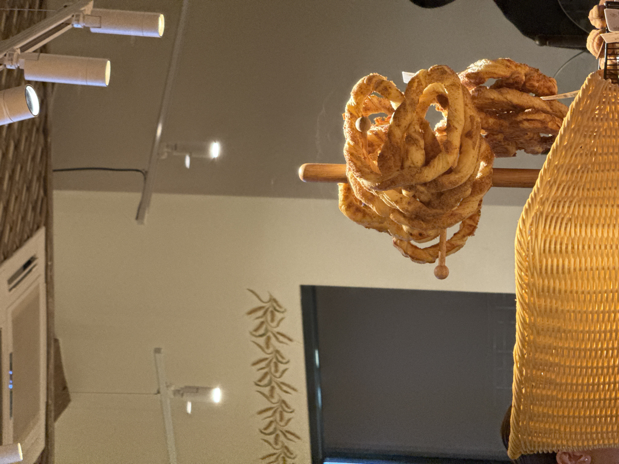 The image shows a commercial interior, likely a bakery or cafe, in Izhevsk, Russia. A bunch of twisted, golden-brown pastries, possibly pretzels or similar baked goods, are suspended from a wooden rod, suggesting they are for sale. A woven, light-colored basket or lampshade is partially visible on the right side, obstructing the view. In the background, there is a wall with a painted vine motif, a window or monitor with a dark screen, and track lighting on the ceiling. The overall lighting is warm and inviting.