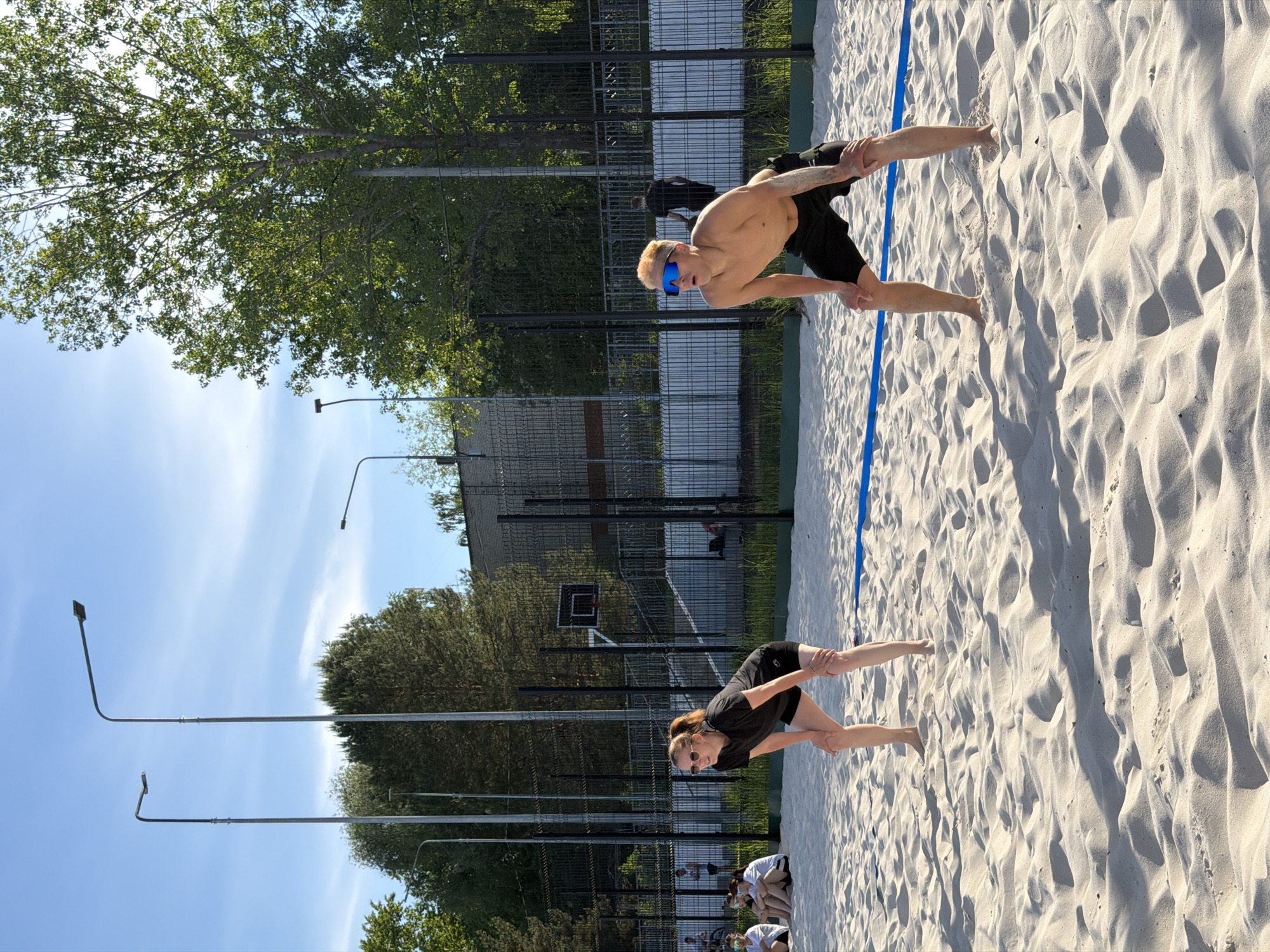 The image depicts two individuals on a sand volleyball court in Chaikovsky, Russia. One is a shirtless man wearing blue sunglasses, and the other is a woman wearing a black shirt. They are both bending over, possibly stretching or warming up, facing away from each other on opposite sides of a blue boundary line. The court is surrounded by a dark green fence and trees. In the background, there are lampposts and buildings, suggesting an urban or recreational area. Other individuals are visible in the lower part of the image, sitting or standing in the background. The sky is blue with light clouds.