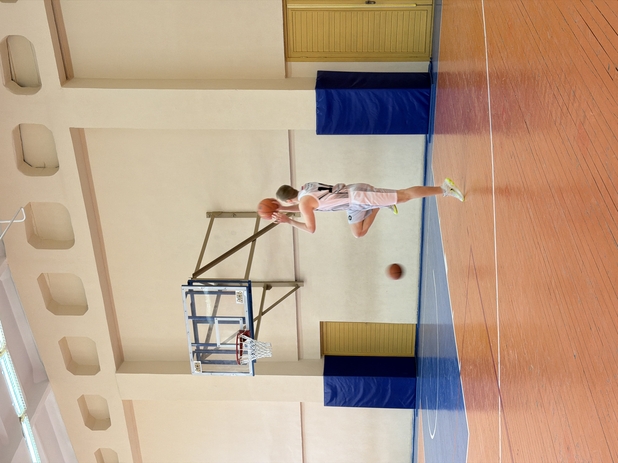 The image depicts a basketball player in mid-air, holding a basketball with both hands, presumably about to shoot. The player is wearing a white and pink jersey with the number 11 and white and grey shorts with the number 0. A second basketball is bouncing on the wooden basketball court. The setting is a gymnasium with light beige walls and a wooden floor, typical of an indoor basketball court. The basketball backboard and hoop are visible. Blue padding is attached to the wall near the backboards. The location is most likely in the city of Chaikovsky, Russia.
