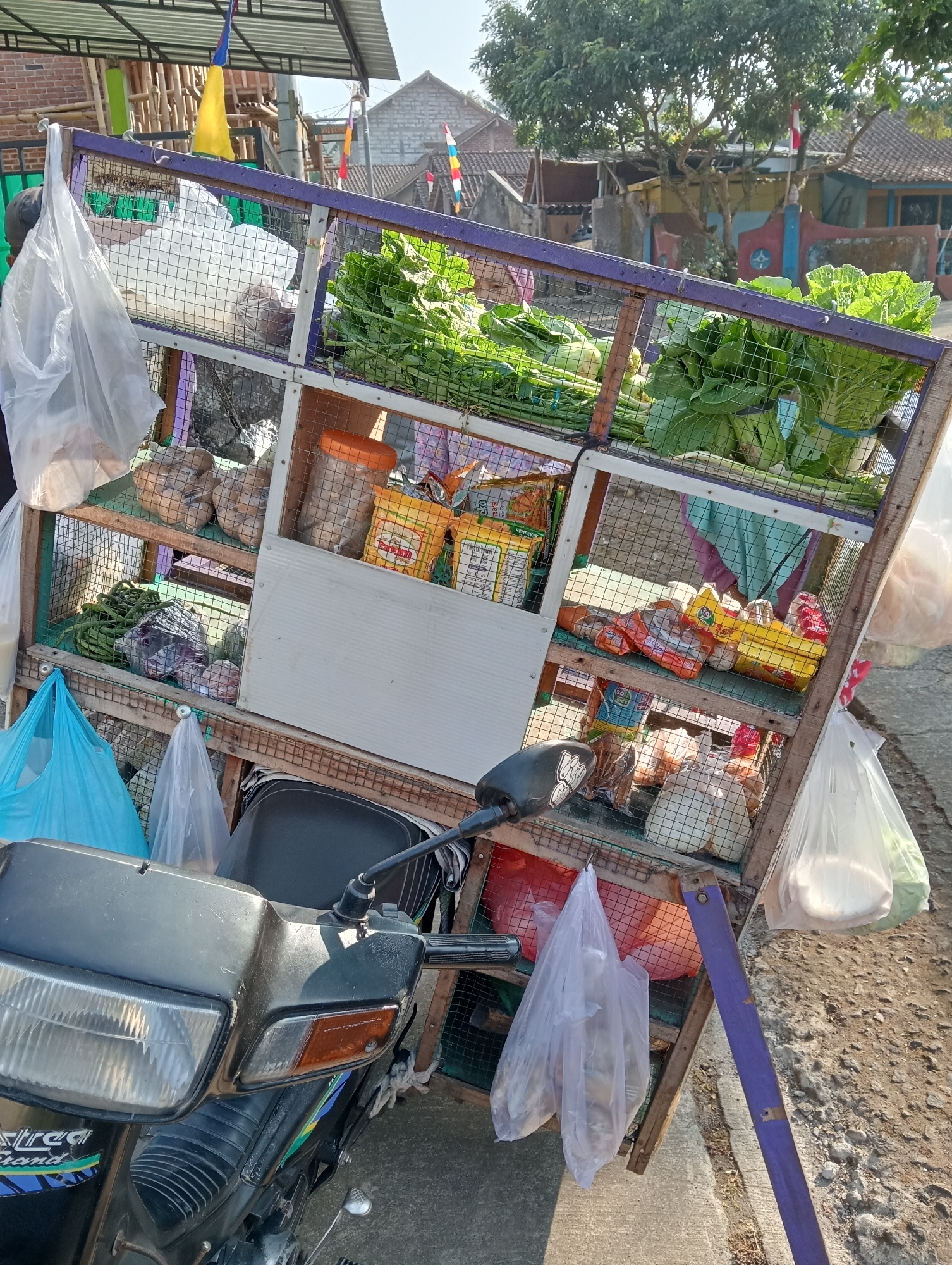 The image depicts a street scene in Krajan Kidul, Indonesia, featuring a mobile market stall mounted on a motorcycle. The stall is constructed from wood and wire mesh, displaying a variety of goods. Prominently featured are large bundles of green leafy vegetables, likely for sale, arranged on the top shelves. Below these, various packaged food items, including what appear to be snacks and dry goods in colorful packaging, are visible. Bags of produce and other items hang from hooks on the stall. The motorcycle, a dark-colored scooter, is positioned in the foreground, serving as the base for the market stall. The background shows residential buildings and trees, indicating a typical village or small town environment. The lighting suggests it is daytime, with sunlight casting shadows. The scene captures a common mode of commerce in many parts of Indonesia, where vendors utilize motorcycles to transport and sell their goods.