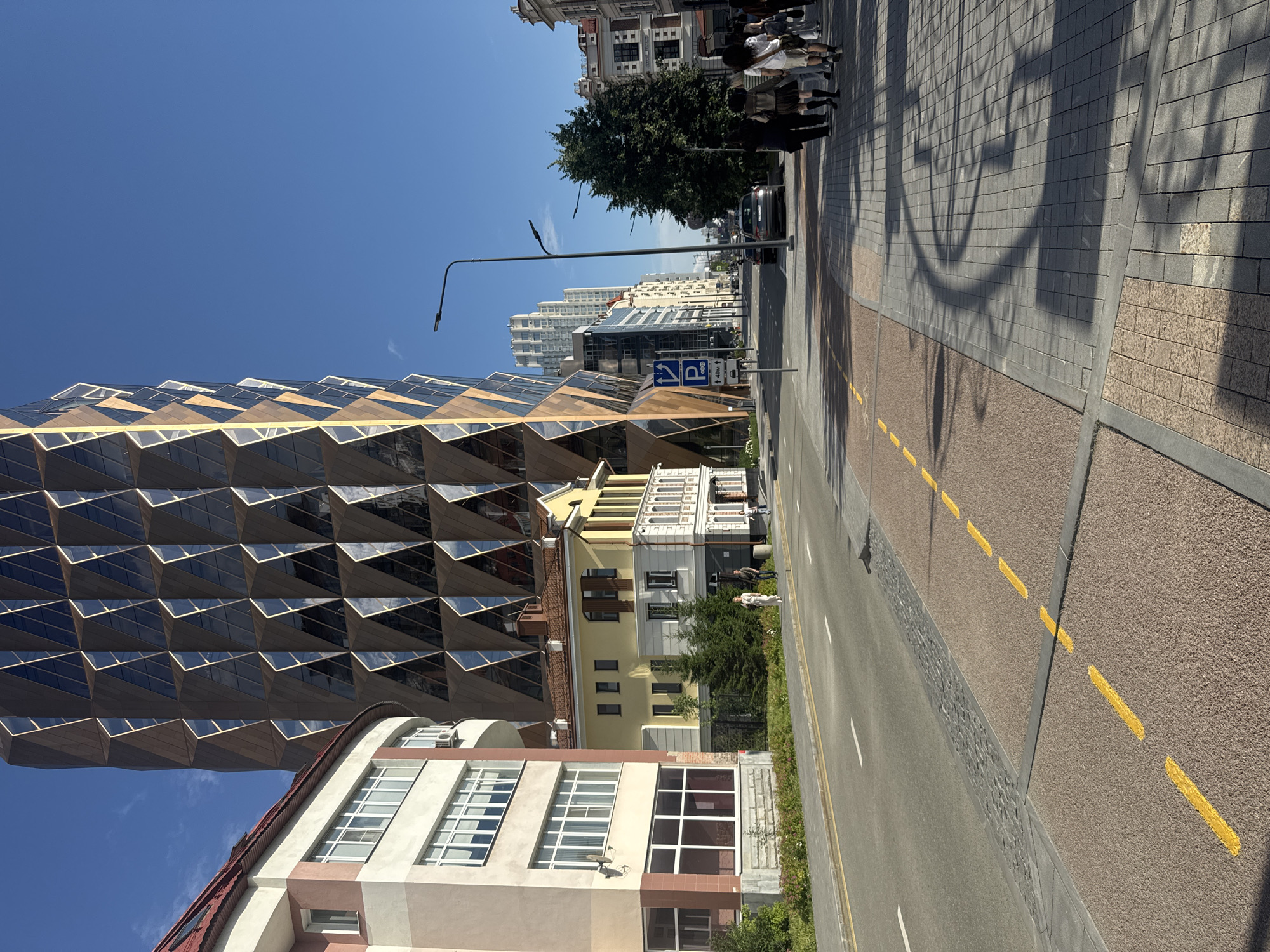 The image depicts a street scene in Yekaterinburg, Russia, under a clear blue sky. In the foreground, a paved road runs from left to right, with a bike lane marked by yellow dashed lines. A sidewalk with a patterned brick surface runs parallel to the road on the right.
Dominating the left side of the frame is a modern, angular building with a facade composed of repeating diamond-shaped glass panels, reflecting the sunlight. To the left of this building is a more traditional, multi-story structure with a reddish roof and light-colored walls.
Further down the street, other buildings of varying architectural styles can be seen, including a yellow building with a red-tiled roof and a row of taller, modern high-rise buildings in the distance. A few vehicles are present on the road, and several people are walking on the sidewalk. A street lamp stands on the right side of the sidewalk. The scene suggests an urban environment with a mix of contemporary and older architecture.