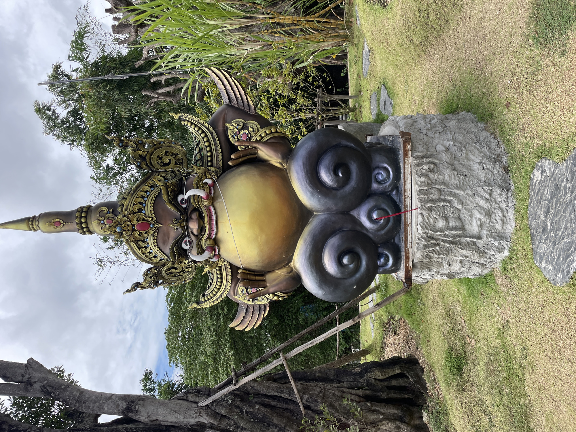 The image shows a large, ornate statue of a mythical creature in an outdoor setting. The statue is golden and dark brown, with a large, round belly and elaborate headdress. It is seated on a dark, swirling cloud-like base, which is placed on a rough, grey stone pedestal. The statue is located in a garden area with lush green grass, a large tree on the left, and tall reeds or sugar cane on the right. A rustic wooden ladder leans against the tree. In front of the statue, on the pedestal, is a small, red incense stick. The background consists of trees and a cloudy sky. The scene appears to be in a public park or garden in the Bo Win Subdistrict, Thailand.