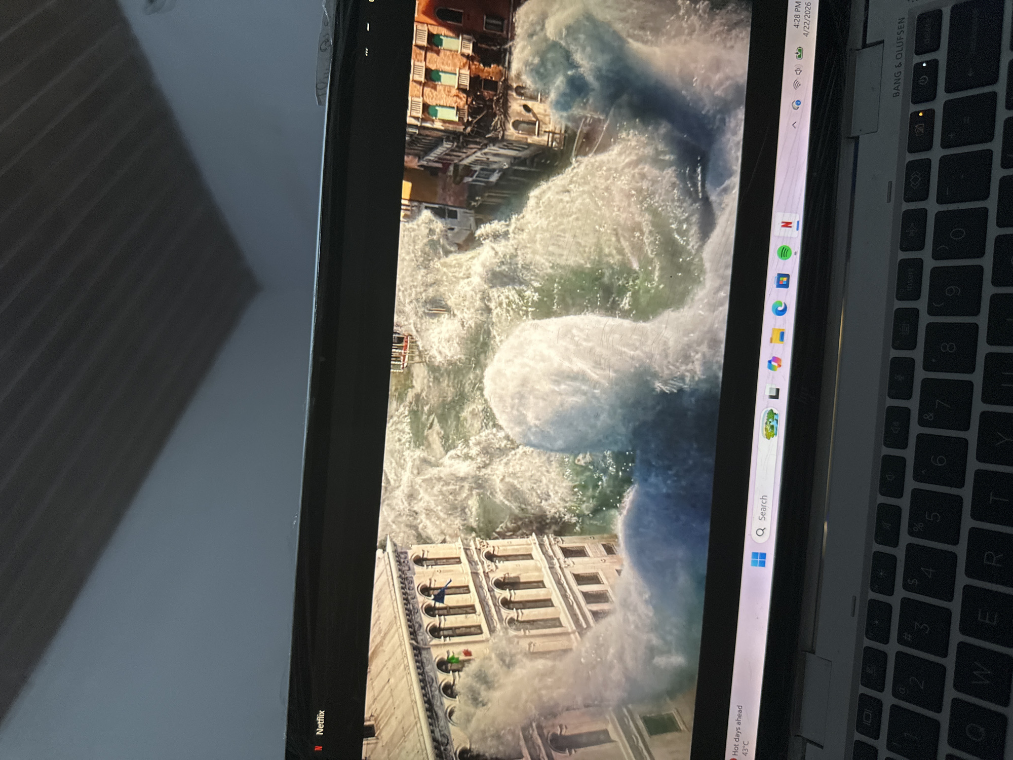 The image captures a laptop screen displaying a dramatic scene of large, turbulent waves engulfing multi-story buildings, suggesting a flood or tsunami in a densely built area. The buildings, some with reddish-brown facades and balconies, appear to be partially submerged or under assault by the water.
A section of the laptop's keyboard, bearing "BANG & OLUFSEN" branding, is visible on the right side of the image. The laptop's operating system interface is discernible, showing the time as "4:28 PM" and the date as "4/22/2026". A weather forecast indicating "Hot days ahead" and a temperature of "43°C" is also visible. This temperature aligns with the provided location context of Kirana, Nigeria, where the photo was taken. Application icons, including Netflix (where the content is likely streaming from), Spotify, and Microsoft Edge, are present on the taskbar. Reflections of internal surroundings, possibly blinds, can be seen on the left side of the screen. The scene on the screen evokes a powerful sense of catastrophe and destruction.