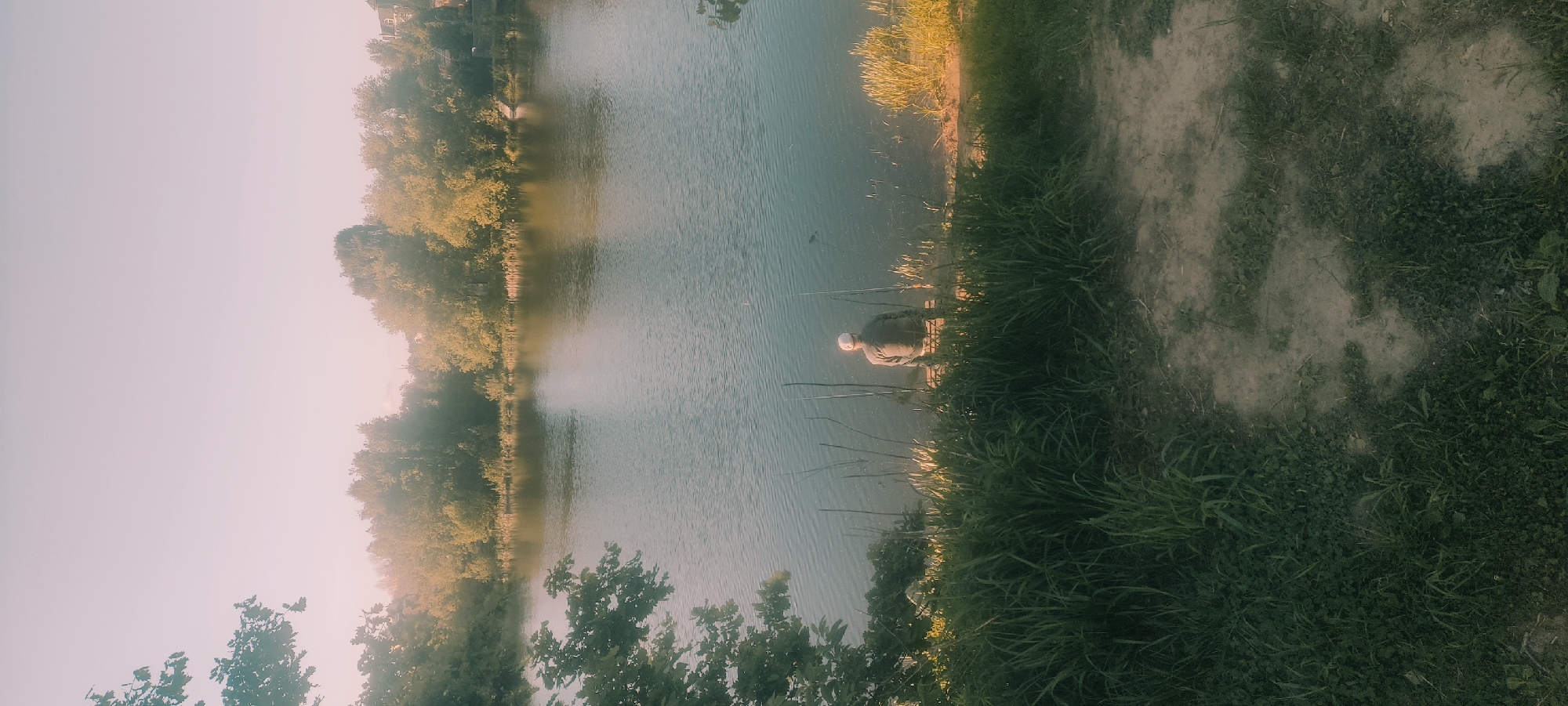 A person is fishing in a lake in Fryazino, Russia. The lake is surrounded by trees and vegetation. The person is sitting on the edge of the lake, holding a fishing rod.