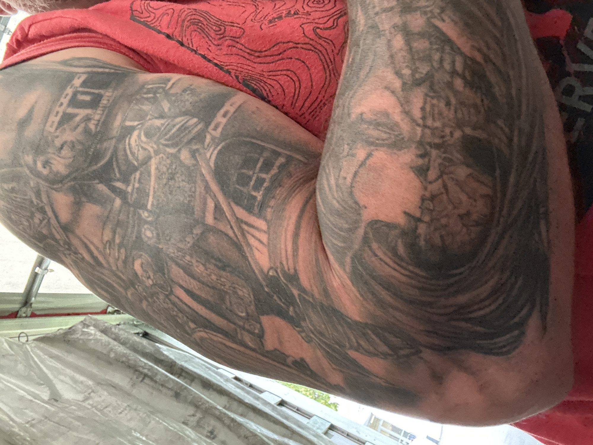 The image shows a close-up of a person's tattooed arm and shoulder, with part of a red t-shirt visible. The tattoo appears to be a detailed, black and grey depiction of a scene that includes a figure in armor, a building with windows, and swirling wave-like patterns. The person is wearing a red t-shirt with a pattern that resembles topographic lines. The background is out of focus but suggests an outdoor environment with some kind of structure and possibly a tent or tarp. There is no discernible connection to Dornbirn, Austria.