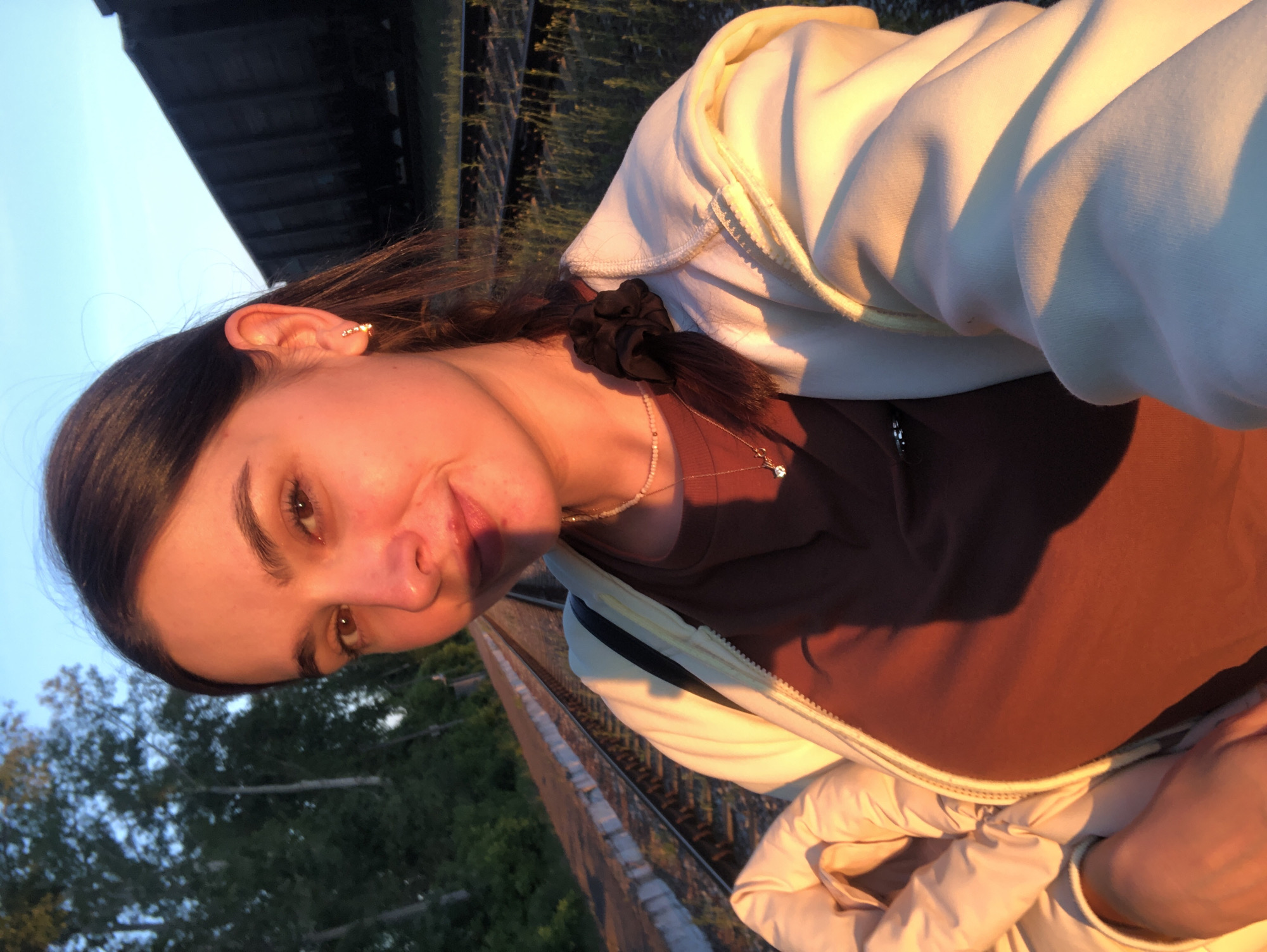 A young woman is depicted in a selfie-style portrait, her head tilted to the side, looking directly at the camera with a gentle smile. She has dark hair pulled back, secured by a black scrunchie. Her visible right ear is adorned with a delicate ear cuff or multiple small earrings. She is wearing a brown or rust-colored t-shirt under a cream or white hooded sweatshirt.
The setting is outdoors, bathed in warm, golden light characteristic of late afternoon or early evening (golden hour). This light strongly illuminates her right side. In the background, railway tracks extend into the distance, flanked by green foliage and trees. A large, dark structure, possibly the underside of a bridge or overpass, looms against a clear, pale blue sky in the upper left of the frame. The location cues suggest an area near railway infrastructure in Chaikovsky, Russia. There is no visible text in the image.