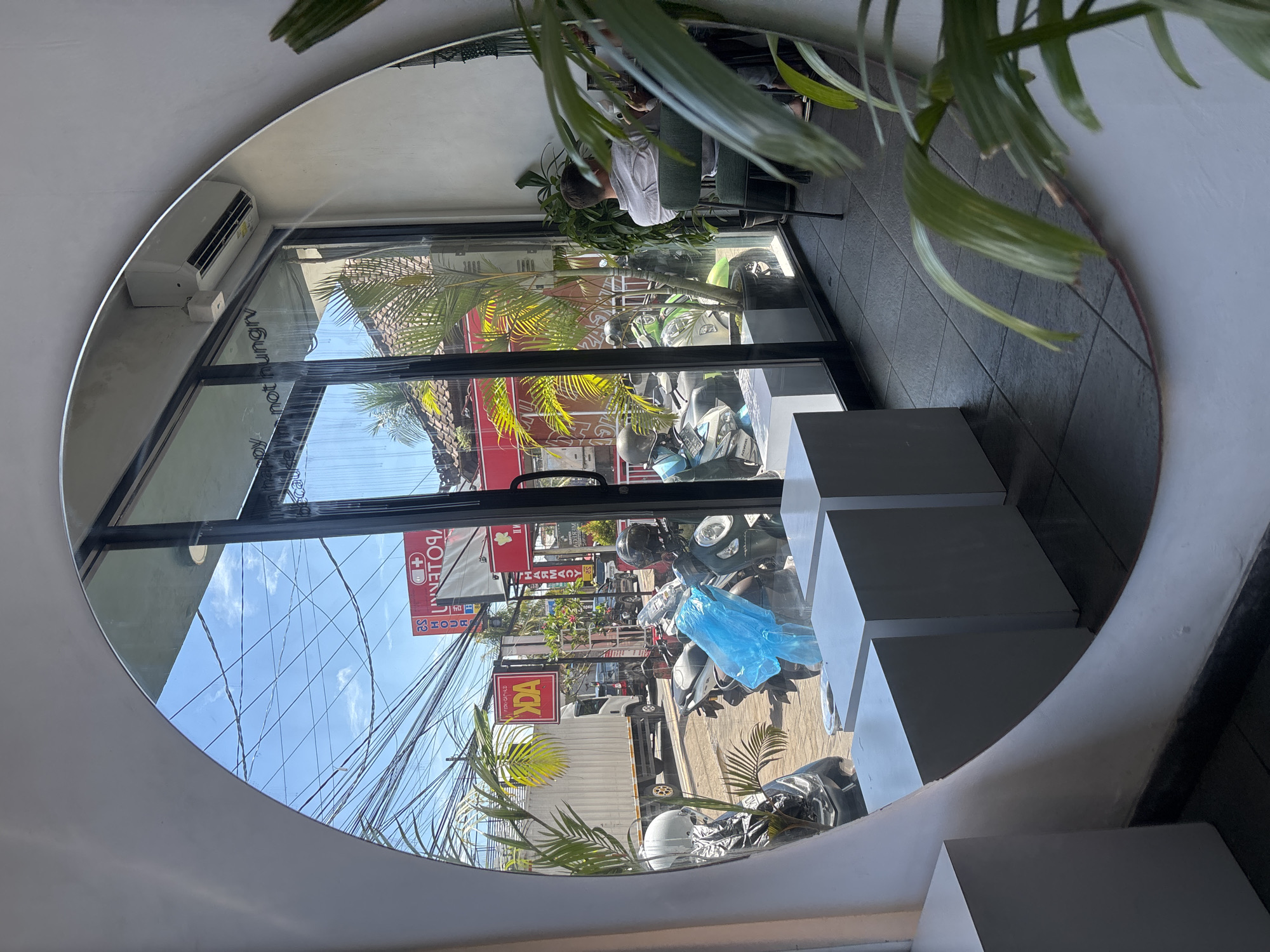 The image is a reflection in a circular mirror, likely inside a building with modern interior design. The reflection shows a street scene in Kuta Selatan, Indonesia.
The street scene includes several motorbikes parked along the side, reflecting the high motorbike use in the area. Signage for a pharmacy ("PHARMACY" and "APOTEK" are visible) is displayed on a red and white sign. Power lines crisscross the sky.
Inside the building, a person is seated, and potted plants are situated near the window/mirror. The interior design features geometric shapes in white. An air conditioning unit is installed on the wall near the top of the window frame.