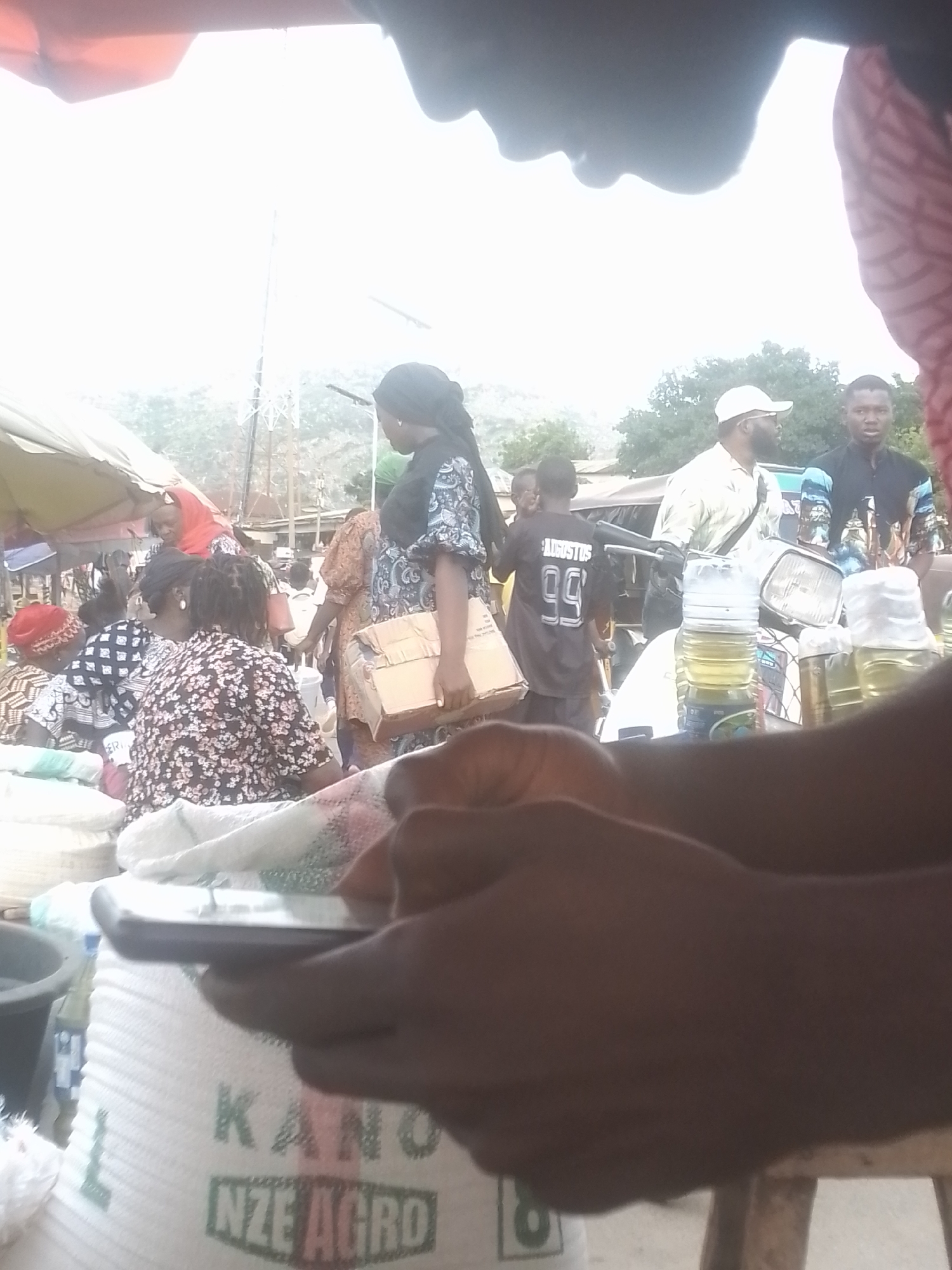 In an outdoor market in Unknown, Unknown, people are seen browsing and shopping. A person's hands are prominently in the foreground, holding a smartphone. Behind the hands, a large white bag with "KANO NZE AGRO" printed on it is visible. In the background, several individuals are gathered around stalls. One woman, wearing a black headscarf and a patterned dress, holds a cardboard box. Another person, a male, is wearing a black t-shirt with "AUGUSTUS 99" on the back. A motorcycle with several bottles of liquid strapped to it is also present in the scene. The market appears to be bustling, with a mix of shoppers and vendors. The environment suggests a warm, possibly humid, day.