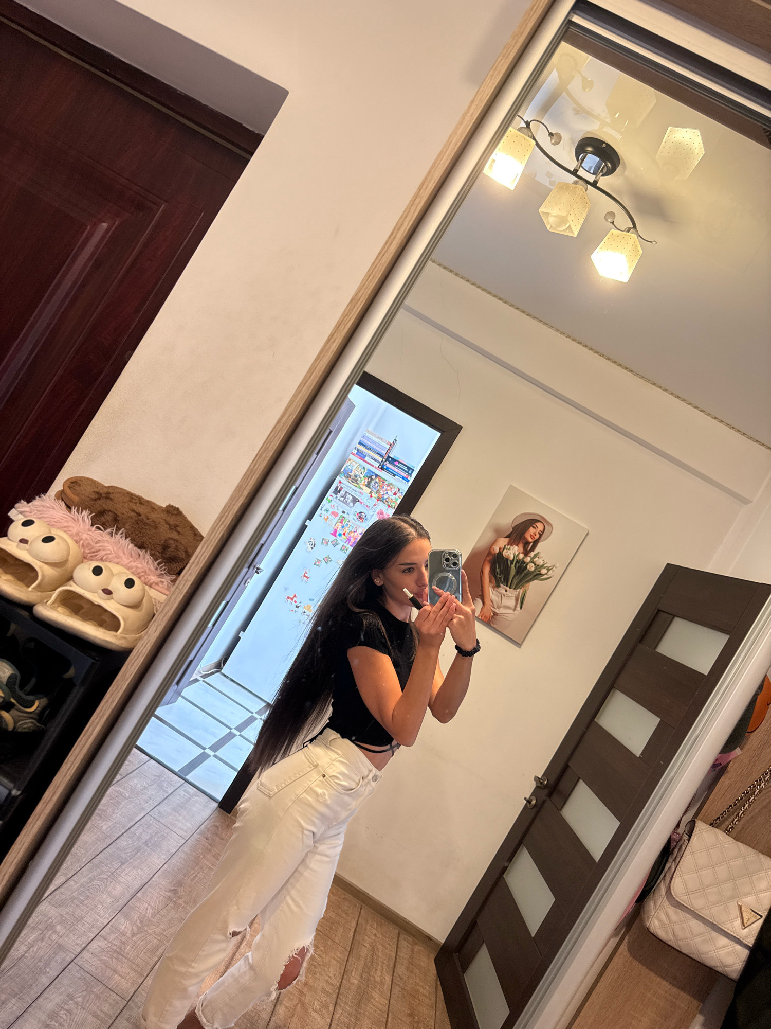 A young woman with long dark hair stands in front of a full-length mirror, taking a selfie with her phone. She is wearing a black crop top and distressed white jeans. In the reflection of the mirror, we can see a hallway with wooden floors, a dark brown door with frosted glass panels, and a framed picture of a woman holding tulips on the wall. A cream-colored quilted handbag with a gold chain strap is visible near the door. To the left of the woman, in the reflection, is a dark wooden door. On a shelf to the left, a pair of novelty slippers shaped like cartoon characters and a fuzzy brown object are visible. The ceiling in the reflection shows a modern light fixture with four square shades. The scene appears to be indoors, likely within an apartment or house.