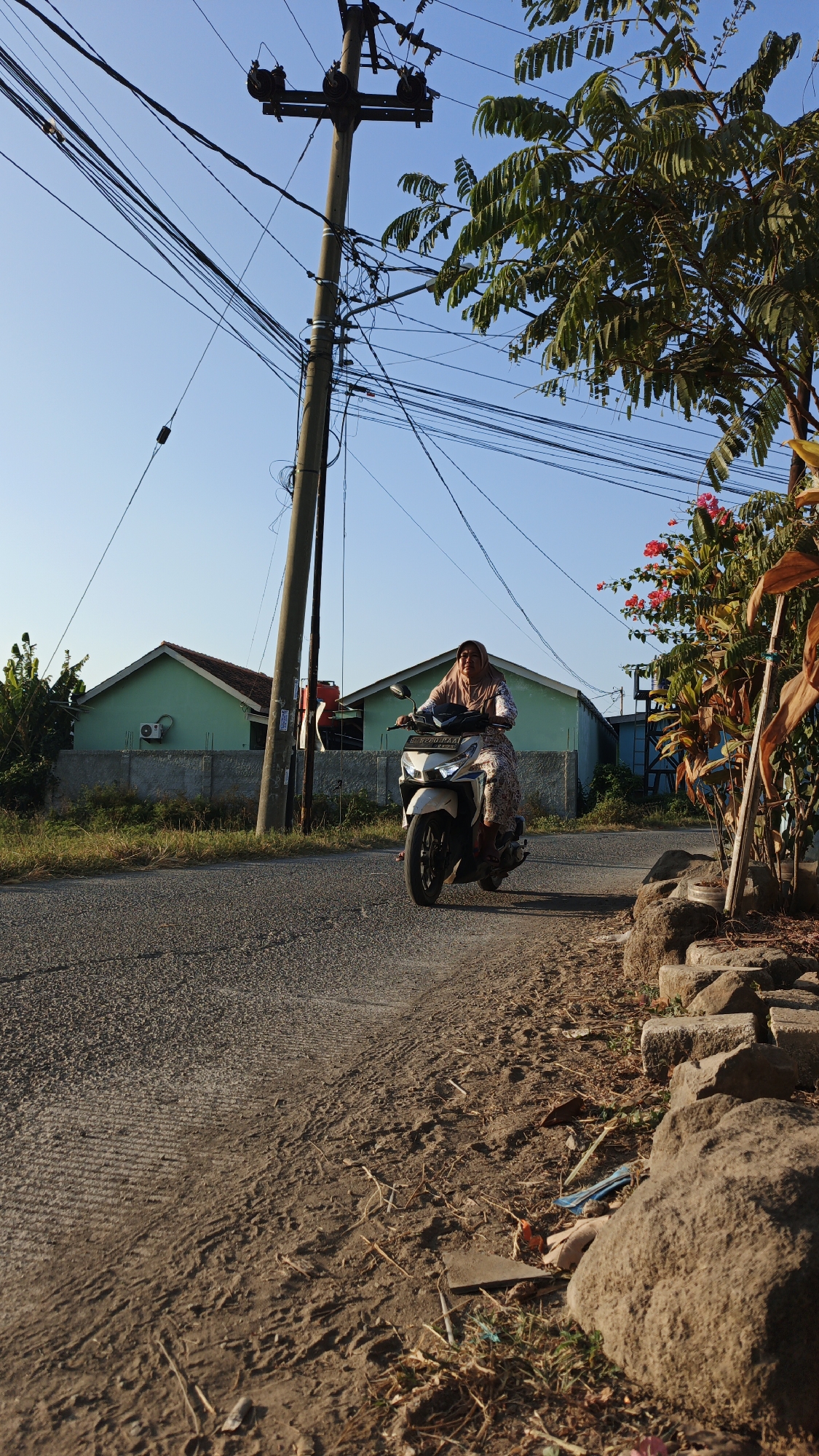 An outdoor, low-angle, medium shot captures a woman riding a white scooter down a paved road in Santing, Indonesia. The woman, wearing a hijab and a patterned dress, is positioned in the center of the frame, her face turned slightly to the left and her gaze directed forward. She is on a scooter with a license plate visible. The road is bordered by a dirt shoulder with scattered rocks and dry grass. In the background, two light green houses with red-tiled roofs are visible, separated by a grey concrete wall. A tall utility pole with numerous black wires crisscrosses the sky behind the houses, and a large tree with green leaves extends from the right side of the frame, partially obscuring a bush with pink flowers. The sky is a clear, pale blue, indicating a sunny day.