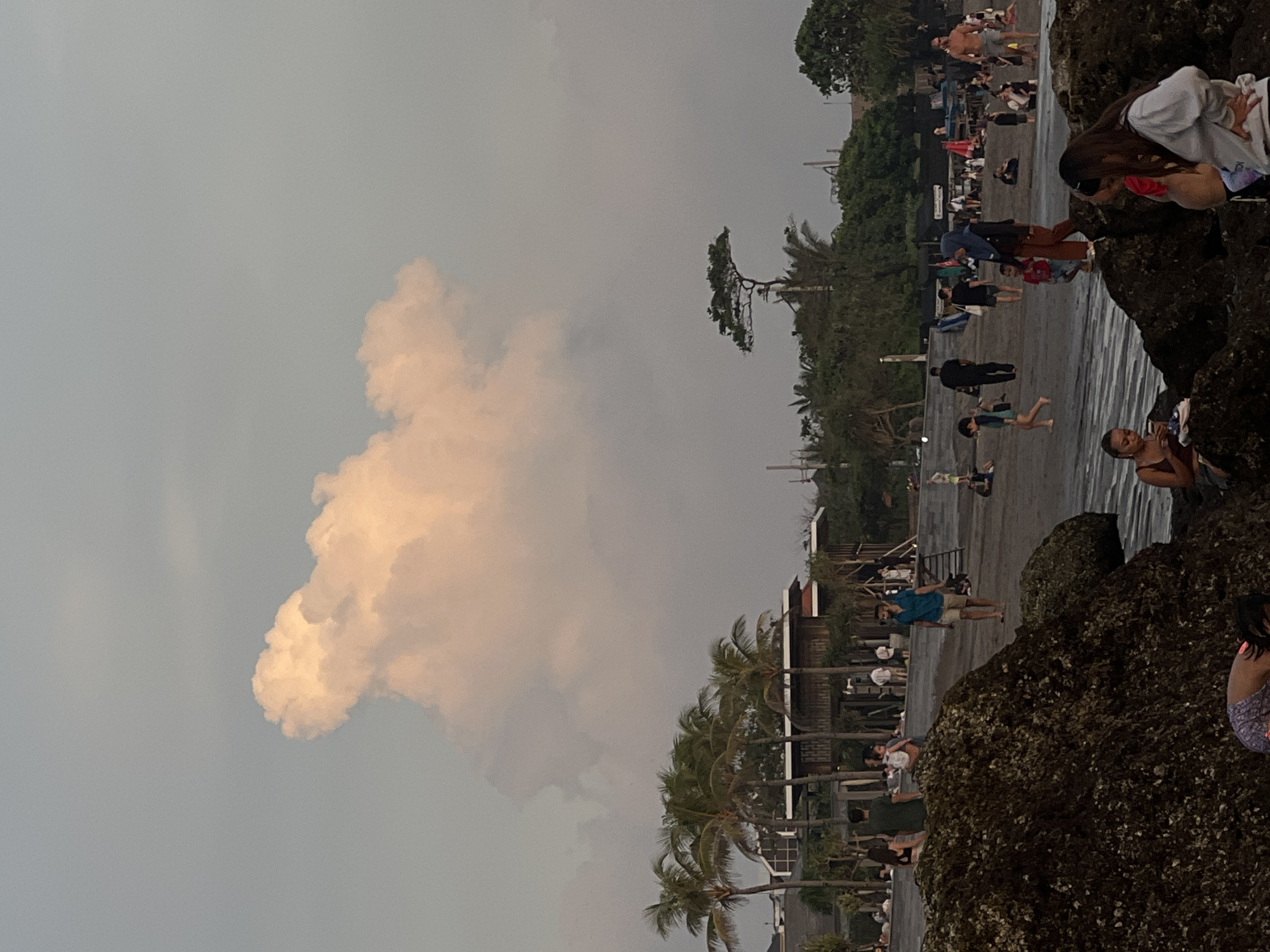 The image shows a beach scene in Canggu, Indonesia. On the right of the image, people are gathered on a beach, some standing on rocks near the water. The beach is crowded with people of various ages. Palm trees are visible along the shoreline. The sky is overcast with a large cloud formation dominating the left side of the image.