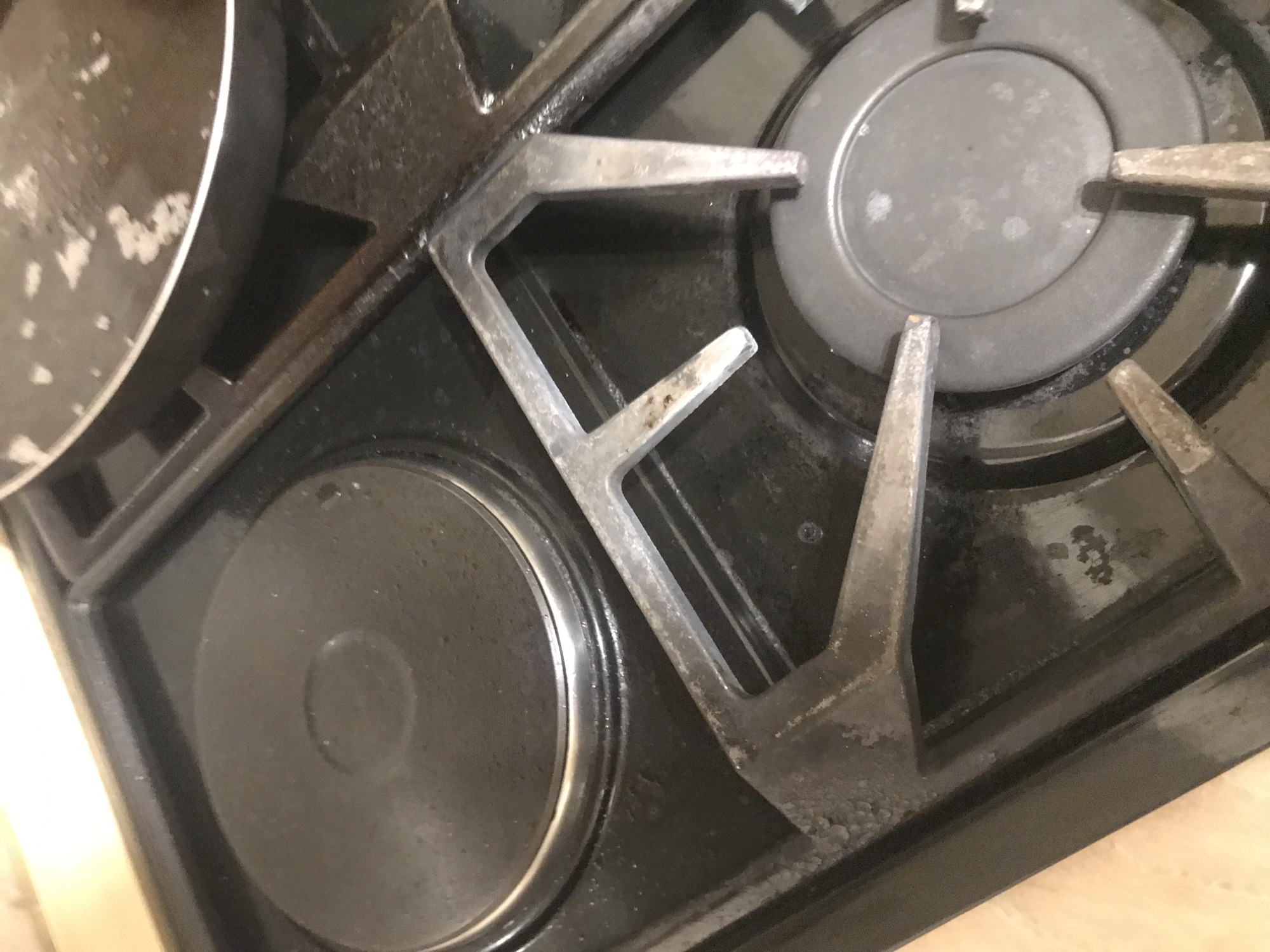 A close-up shot depicts a black gas stovetop. Two burners are visible: one electric coil burner on the left and a gas burner on the right. A metal pan partially obscures the top left of the image. The stovetop and burner grates show signs of use with some staining and residue. The image does not contain identifiable information about Yerevan, Armenia.