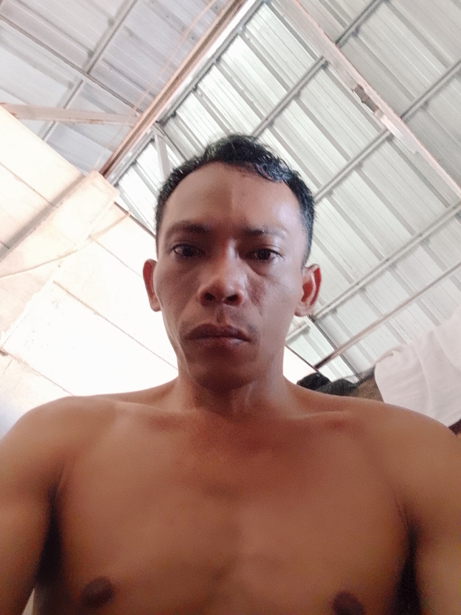 A shirtless man is seen from a low angle against a corrugated metal roof. The man is dark-skinned with short, dark hair. The location is Bukit Ketok, Indonesia.