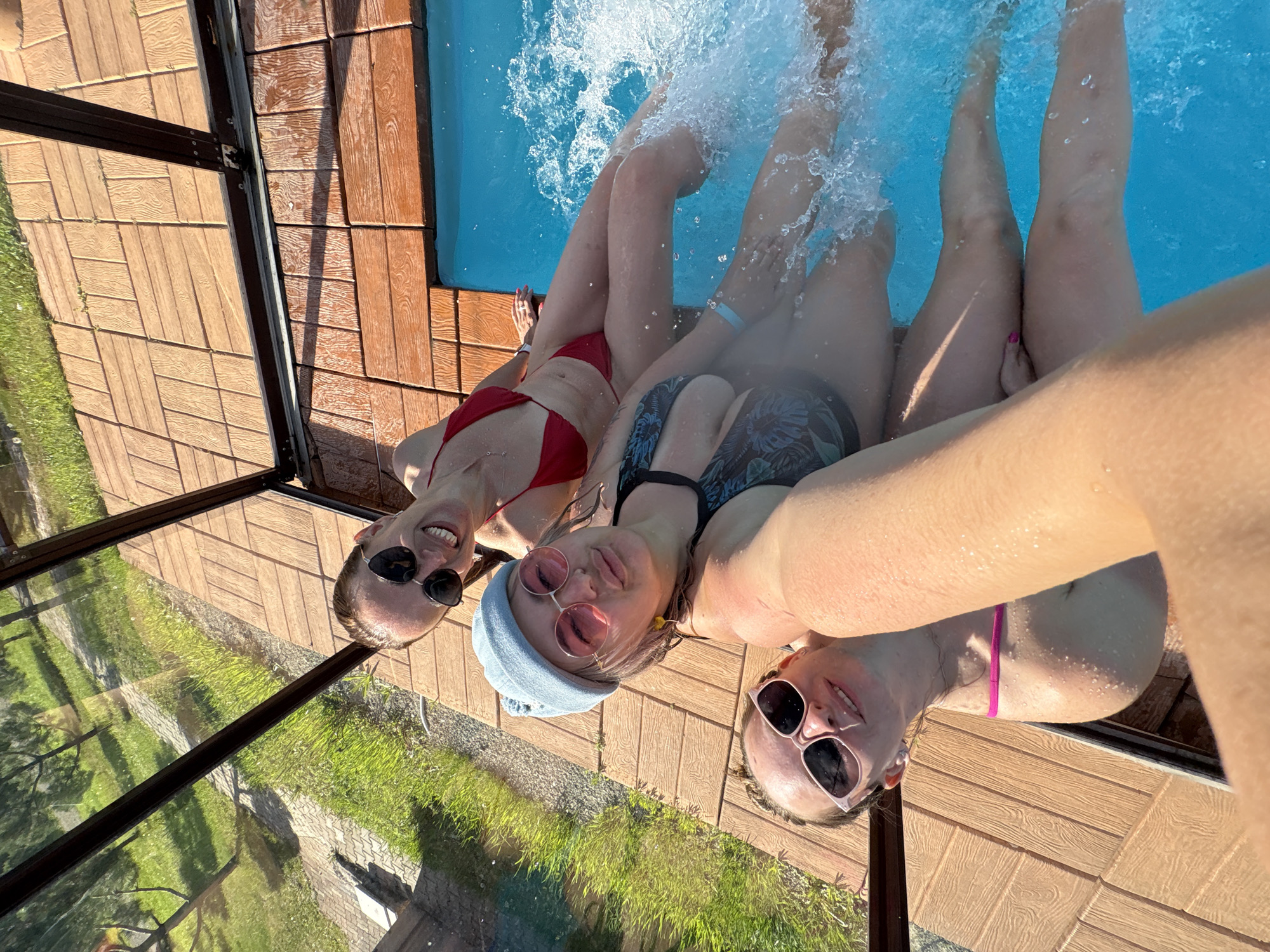 Three young women are posing for a selfie in a swimming pool. The woman in the foreground is wearing a blue beanie and pink sunglasses. The woman in the middle is wearing a red bikini. The woman in the background is wearing a patterned bikini. They are all smiling and appear to be enjoying themselves. The pool is surrounded by wooden decking. The scene takes place outdoors on a sunny day. There is no indication that this location is in Birobidzhan, Russia.