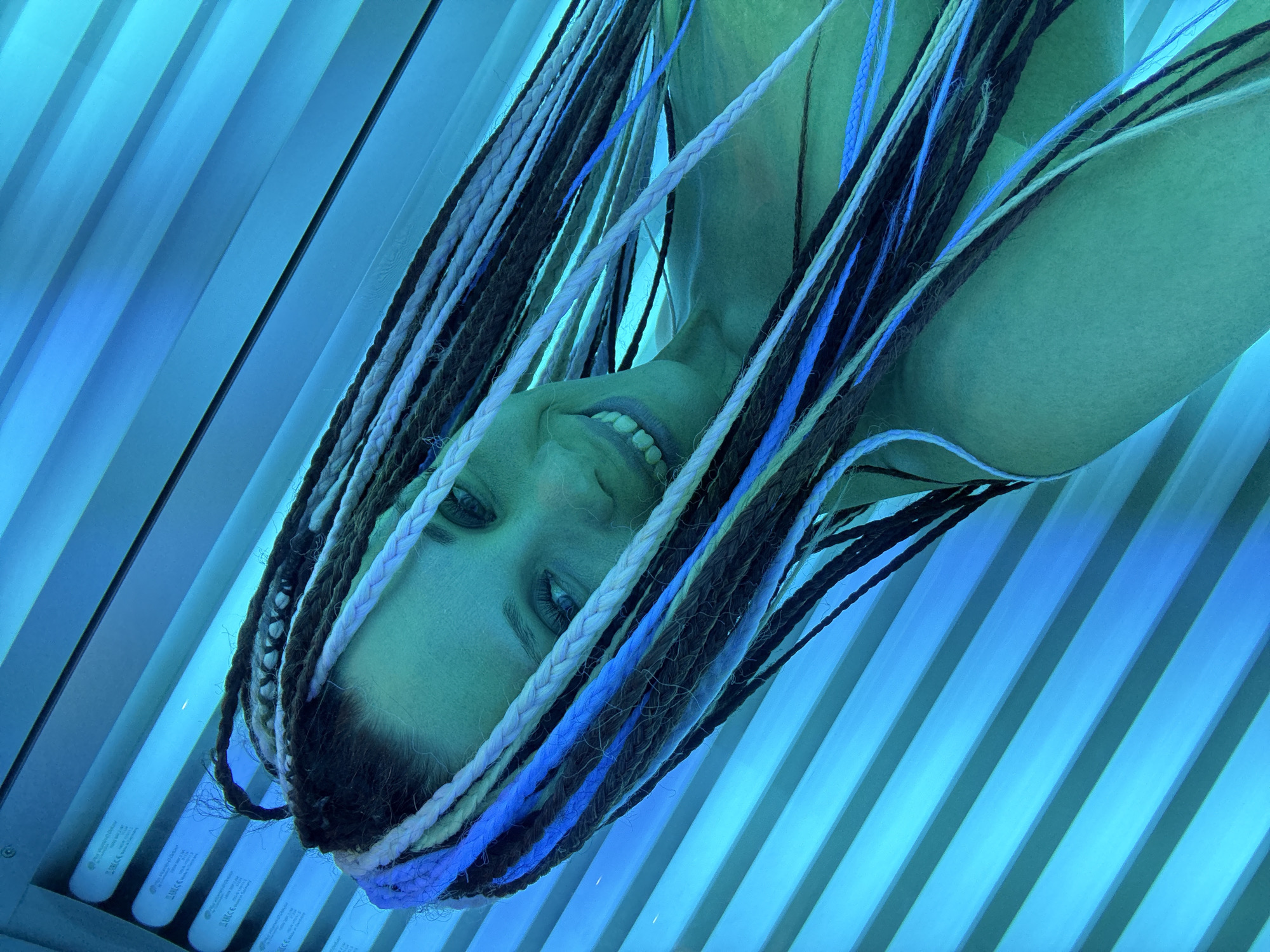 A young adult female is captured in a close-up, low-angle shot, lying inside what appears to be a tanning bed or solarium. She smiles broadly at the camera, showing her upper teeth. Her face and visible skin have a distinct pale green-blue tint due to the vibrant blue light emitted by the fluorescent tubes that surround her. Her hair is styled in long, prominent braids (likely dreadlocks or similar synthetic extensions) in a variety of colors, including shades of brown, black, white, and striking electric blue strands, which fan out around her head.
The setting is characterized by rows of glowing blue fluorescent light tubes, typical of a tanning booth, which provide the primary illumination for the scene. The internal surfaces of the tanning bed reflect this blue light, creating a uniformly cool, blue-green environment. Visible text on some of the light tubes includes phrases like "UV Lamps," "EN 60335-2-27," "made in China," and "CE" markings, indicating product specifications. The woman's expression suggests happiness or amusement, consistent with a casual moment captured during a tanning session in a facility, possibly in Rusanovo, Russia.